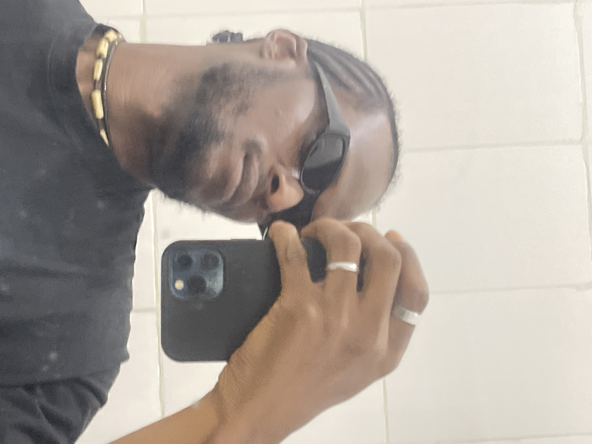 A male individual with dark skin, cornrows, a beard, and wearing dark sunglasses is depicted taking a mirror selfie. He wears a black t-shirt and a necklace featuring light-colored, elongated beads on a dark string. Two silver-colored rings are visible on his right hand, one on the index finger and one on the ring finger. He holds a black smartphone, consistent with an iPhone Pro model due to its visible triple-lens camera array, in his right hand. The background consists of a plain white tiled wall with discernible grout lines, indicating an indoor setting. The specific location in the city of Jos, Nigeria, is not visually verifiable from the image.