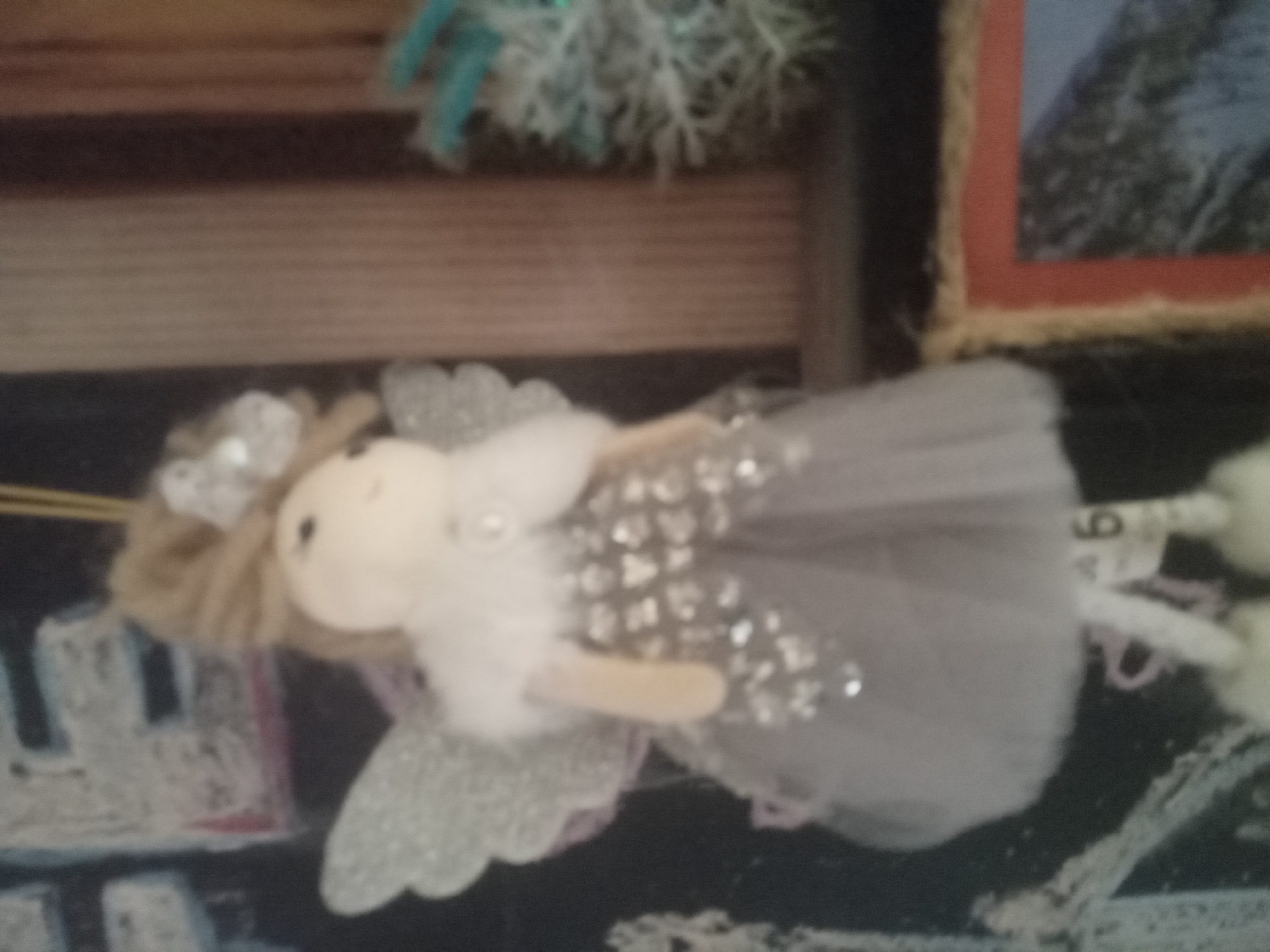 A blurry, close-up, vertical shot features a Christmas ornament of an angel. The angel has light-colored yarn hair and a white fluffy collar. Its dress is light gray with silver embellishments. The angel has white, glittery wings and appears to be hanging from a string. The background is dark and out of focus, with hints of a blackboard or chalk drawings, and a blurred wooden surface. A framed picture is partially visible on the right side of the image, and a blurry evergreen branch with artificial snow is also present.