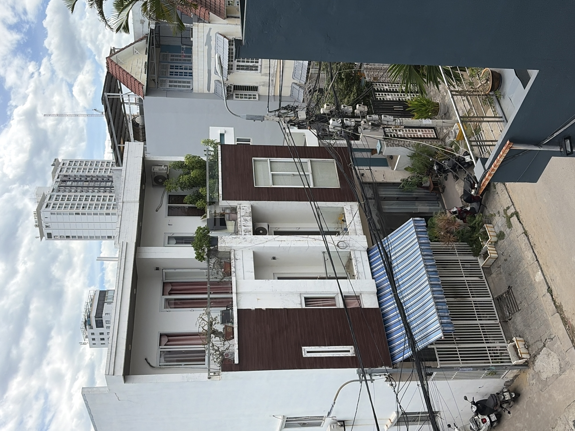 This image depicts a street scene in the city of Đà Nẵng, Vietnam, on a cloudy day. The primary focus is a multi-story building with a white facade and dark brown paneling on the lower section. The building features balconies with metal railings, some adorned with potted plants, and windows with dark red curtains. A blue and white striped awning extends over the ground-level entrance, which is behind a white metal gate. Two scooters are parked on the sidewalk to the left of the building.
In the background, other buildings of varying heights and architectural styles are visible, including a modern, multi-story white building with numerous windows and a taller, slender building with a spire or antenna. Electrical wires and utility poles are prominent features of the urban landscape, crisscrossing the scene. Some of the buildings have air conditioning units mounted on their exteriors. The street is paved and appears to be a typical urban thoroughfare. Several potted plants are strategically placed along the sidewalk and on balconies, adding touches of greenery to the environment. The sky is overcast with visible clouds.