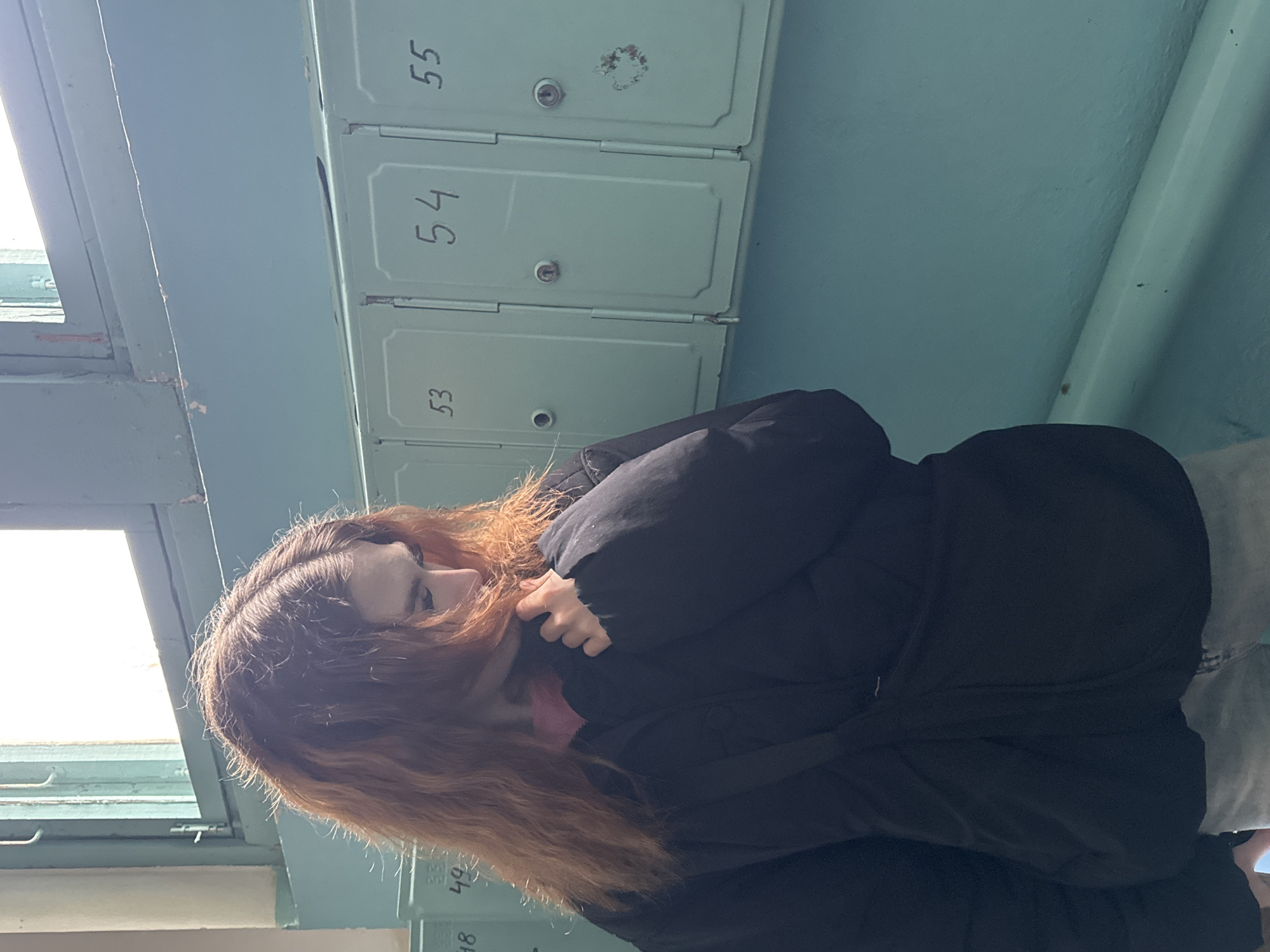 The image, captured in Noviy, Russia, presents a candid, portrait-oriented view of a young person in an indoor hallway or common area. The subject, likely female, is positioned on the right side of the frame, facing left. She has long, wavy reddish-brown hair, illuminated by bright natural light coming from a window to her left. She is wearing a black puffer jacket, with a glimpse of a pink garment visible underneath. Her left hand is raised, appearing to adjust her jacket collar or touch her neck. Her face is partially obscured by her hair and hand, and her gaze is directed downwards or away from the camera, suggesting a pensive or neutral expression.
The background features a light blue wall against which a row of light blue-green metal mailboxes or lockers is mounted horizontally. Three mailboxes are prominently visible, numbered "53," "54," and "55" in black, handwritten text. Some signs of age and wear, including a small rusty spot on mailbox 55, are apparent. Faintly visible below these are parts of other mailboxes with numbers like "48" and "49." A brightly lit window on the far left of the frame indicates that the photo was taken during daylight hours.