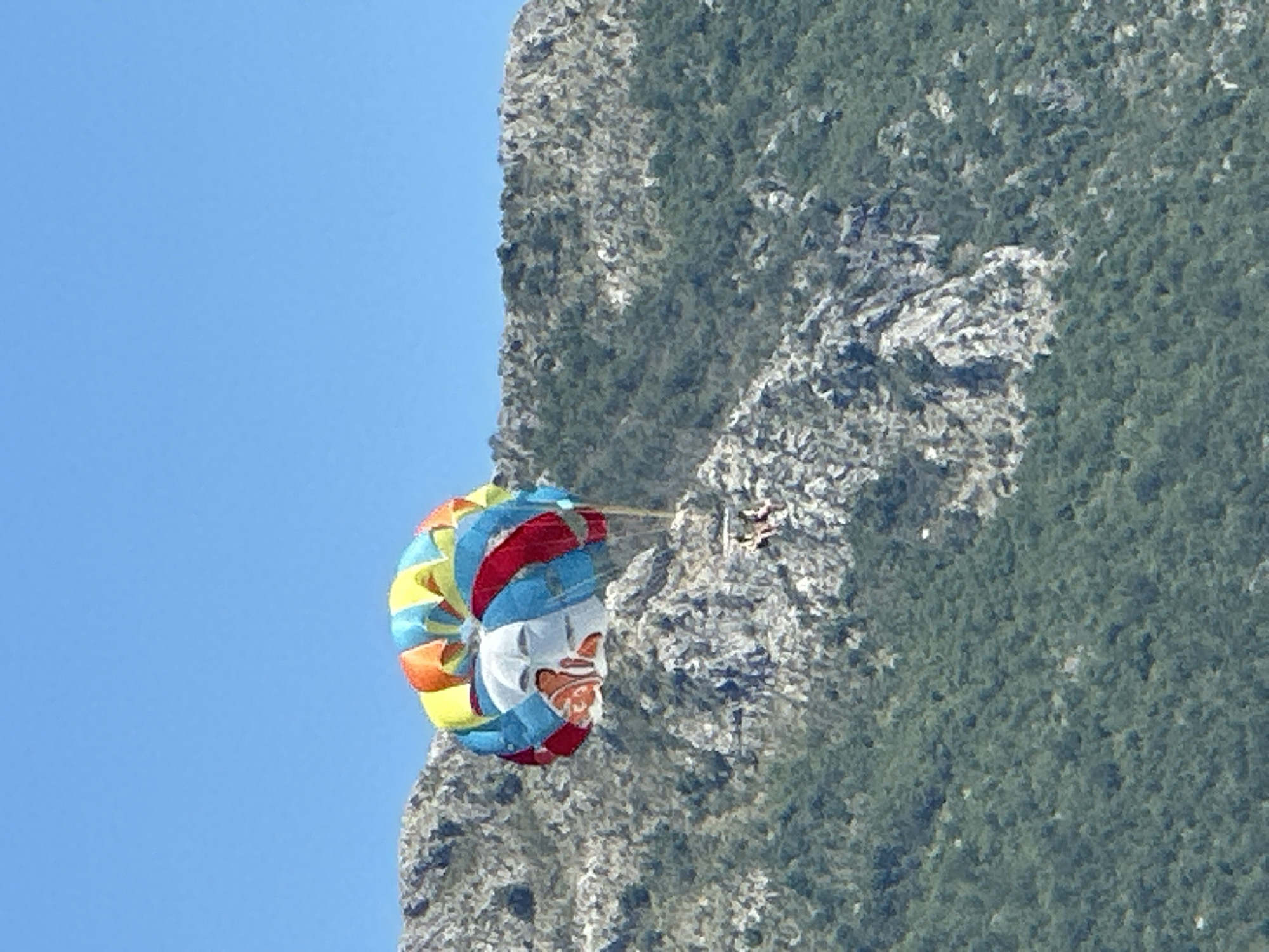 The image captures a parasailor suspended in the sky against a backdrop of a steep, rocky mountainside covered in dense green foliage. The parasail, a vibrant mix of red, blue, yellow, and orange, is inflated and appears to be in mid-flight. Below the parasail, two figures are visible, presumably the individuals being towed. The mountain itself is characterized by rugged, grey rock formations interspersed with dark green vegetation, suggesting a natural, possibly undeveloped area. The sky is a clear, bright blue, indicating good weather conditions. While the image is taken outdoors, the specific location in Boreti, Montenegro, cannot be confirmed from the visual information alone.