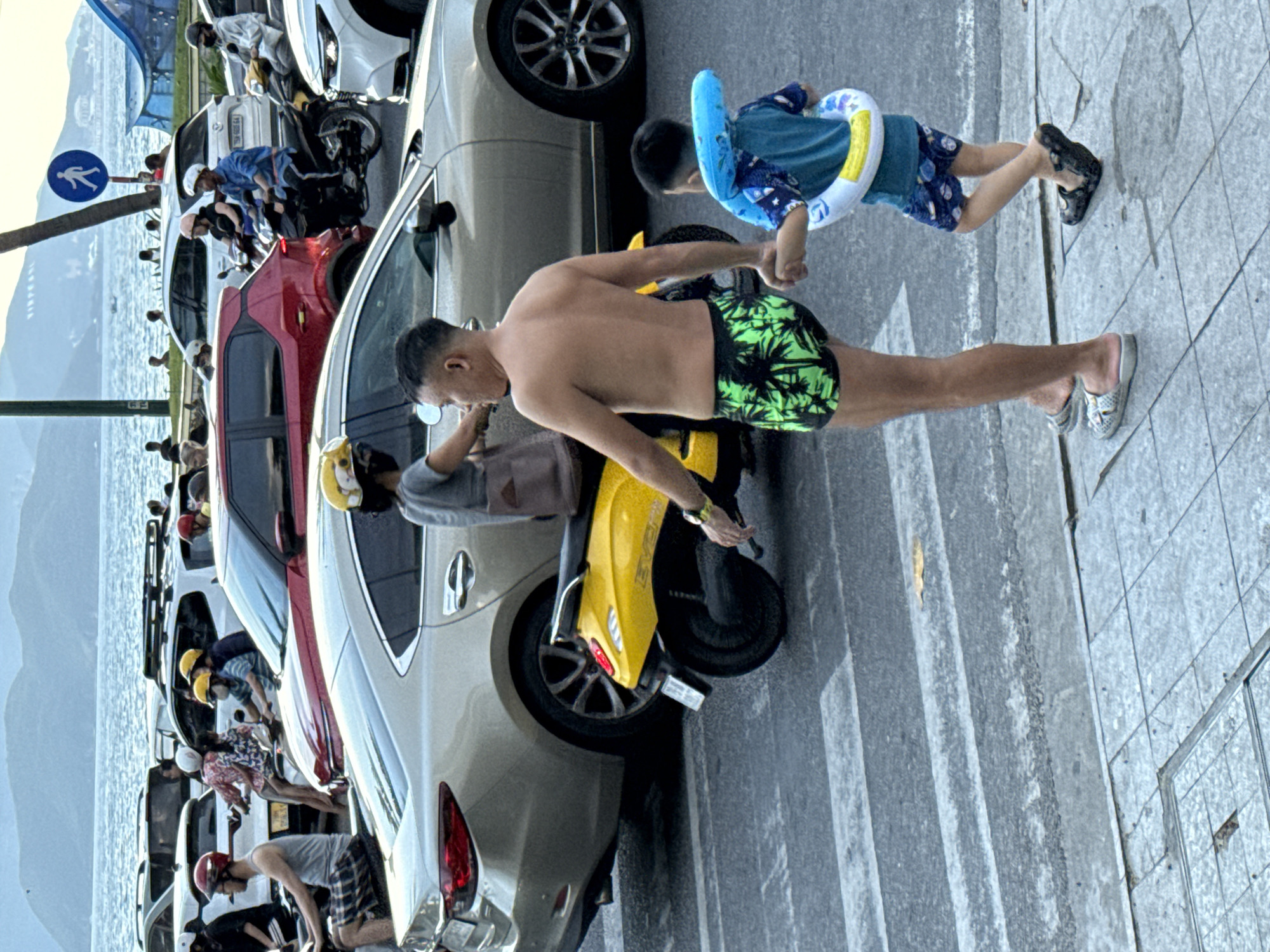In Nha Trang, Vietnam, a man and a child are on a sidewalk next to parked cars and a body of water. The man, shirtless and wearing green palm-print swim trunks, appears to be interacting with a yellow scooter. The child, wearing a blue shirt and patterned shorts, has a blue inflatable ring around their torso. Behind them, several cars are parked in a row, with a red car in the foreground. The scene suggests a warm, possibly sunny day, given the attire of the people. The presence of the water and the beach-like attire hints at a coastal location. The people's actions suggest preparation for or returning from a swim. No visible text is present in the image.