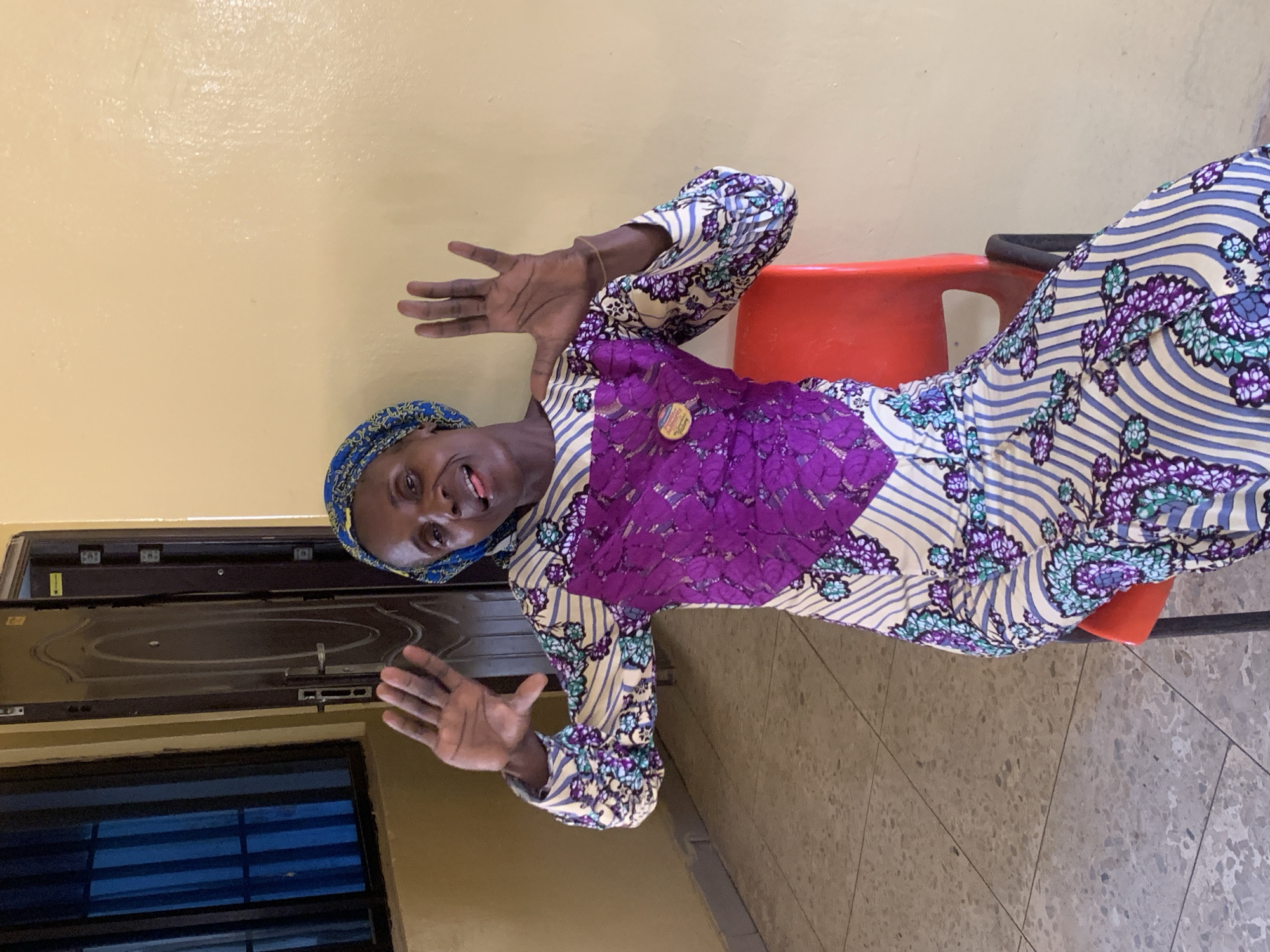A woman wearing a blue head covering and a patterned dress with purple lace detail is sitting on a red chair indoors. She is gesturing with both hands, palms forward, smiling at the camera. Behind her is a beige wall, a dark brown door, and a window with blue blinds. The floor is tiled in a speckled pattern. This image was taken in Apata Yakuba, Nigeria.