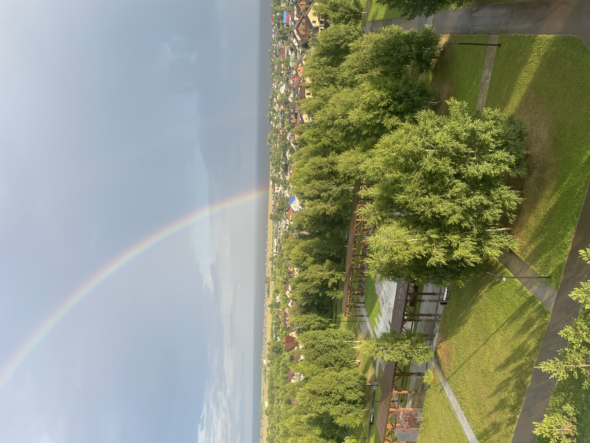 The image shows an aerial view of a park-like area in Laishevo, Russia, with a rainbow arcing across a cloudy sky. In the foreground, lush green trees with birch-like bark dominate the scene, interspersed with grassy areas and paved pathways. Several wooden gazebos or pergolas with open sides are scattered throughout the park. Paved walkways, some wet from recent rain, wind through the landscape, connecting different sections of the park and leading towards a residential area in the middle ground. This residential area consists of numerous houses with pitched roofs of various colors, suggesting a suburban or village setting. Beyond the houses, a body of water, likely a lake or river, stretches to the horizon, where it meets a hazy sky. The overall impression is a peaceful, green landscape under a dramatic sky, indicative of post-rain weather.