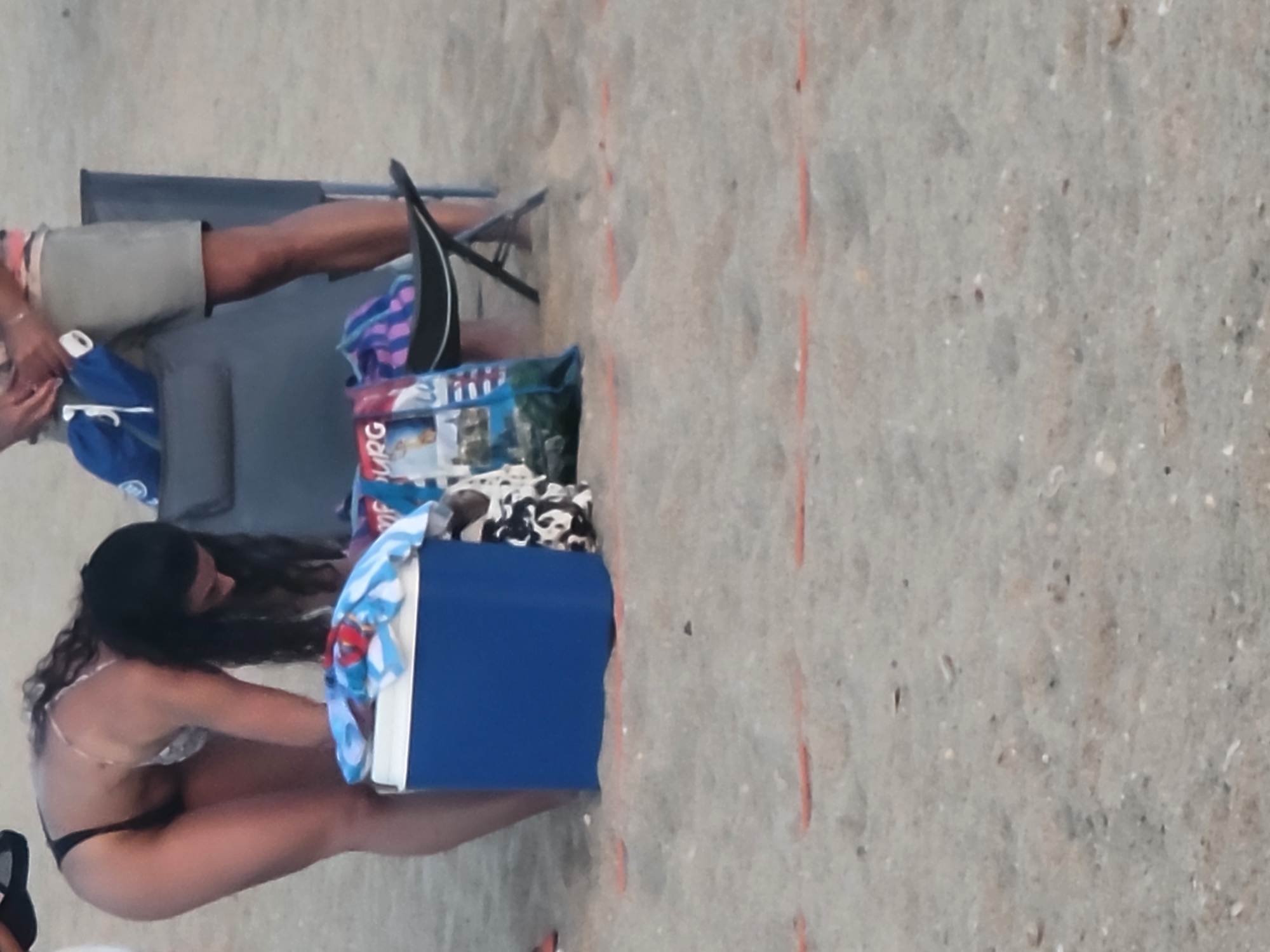 The image shows a beach scene in Quarteira, Portugal. In the foreground, a blue cooler with a white lid sits on the sand, with a blue and white striped towel draped over it. To the right of the cooler, a beach bag with colorful patterns is visible. A woman wearing a dark bikini and a white and patterned top is bending over the cooler. Her dark hair is long and wavy. Behind her, a folding chair is set up. A person wearing khaki shorts and a striped shirt is partially visible sitting in the chair. The ground is covered in sand, with some small, scattered shells and pebbles. In the background, the environment appears to be a beach with the sea possibly out of frame. The overall lighting suggests it is late afternoon or early evening.