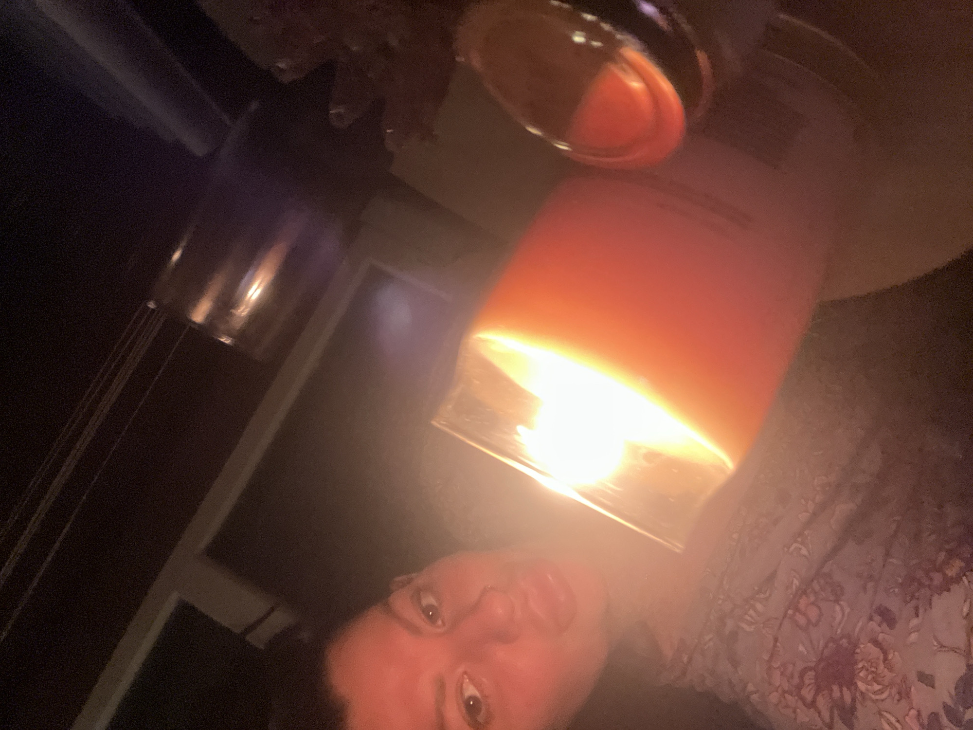 The image shows a close-up, low-light selfie of a person's face and upper body, illuminated by the glow of a large, orange candle in a glass jar. The person, likely female, has their head tilted and is looking towards the camera. They are wearing a floral-patterned garment. The background is dark, with some indistinct objects including what appears to be a wine glass or a similar vessel with a reflective surface. The overall ambiance is intimate and dimly lit. There are no identifiable landmarks or context to suggest a specific location within Plovdiv, Bulgaria.