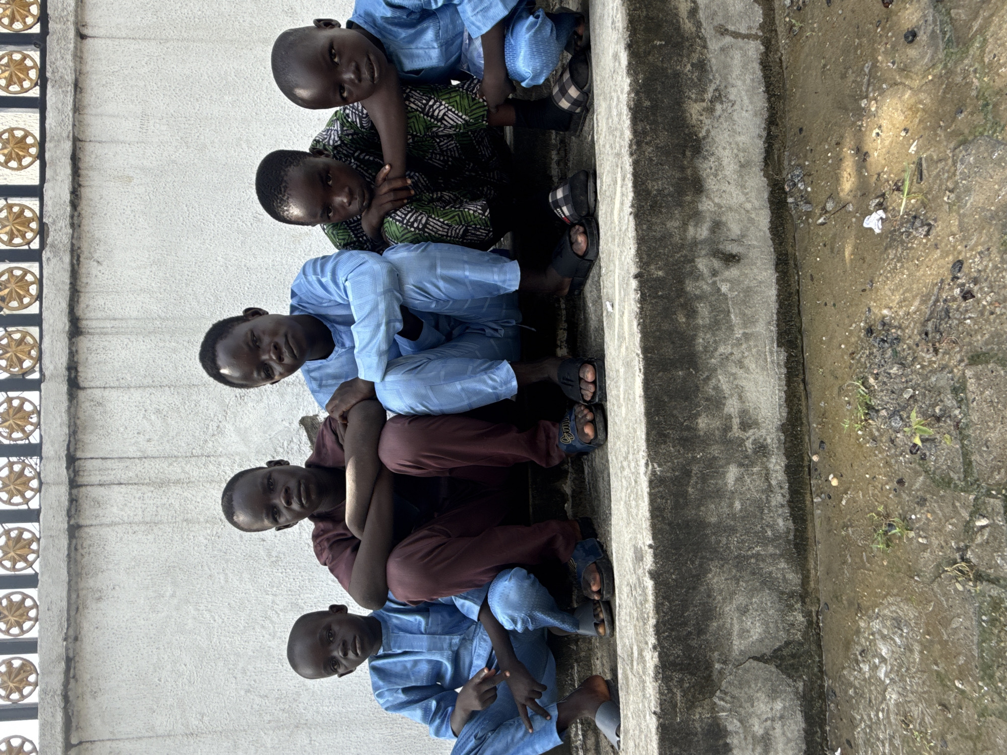 Six boys sit in a row on a concrete ledge in Ado, Nigeria. The ledge is adjacent to a stone wall with a decorative metal fixture. The ground is wet, with small stones and tufts of grass. The boys are wearing a mix of clothing; blue button-down shirts and matching pants, a maroon-colored outfit, and a green patterned shirt. Most are wearing sandals. They are all looking toward the camera.