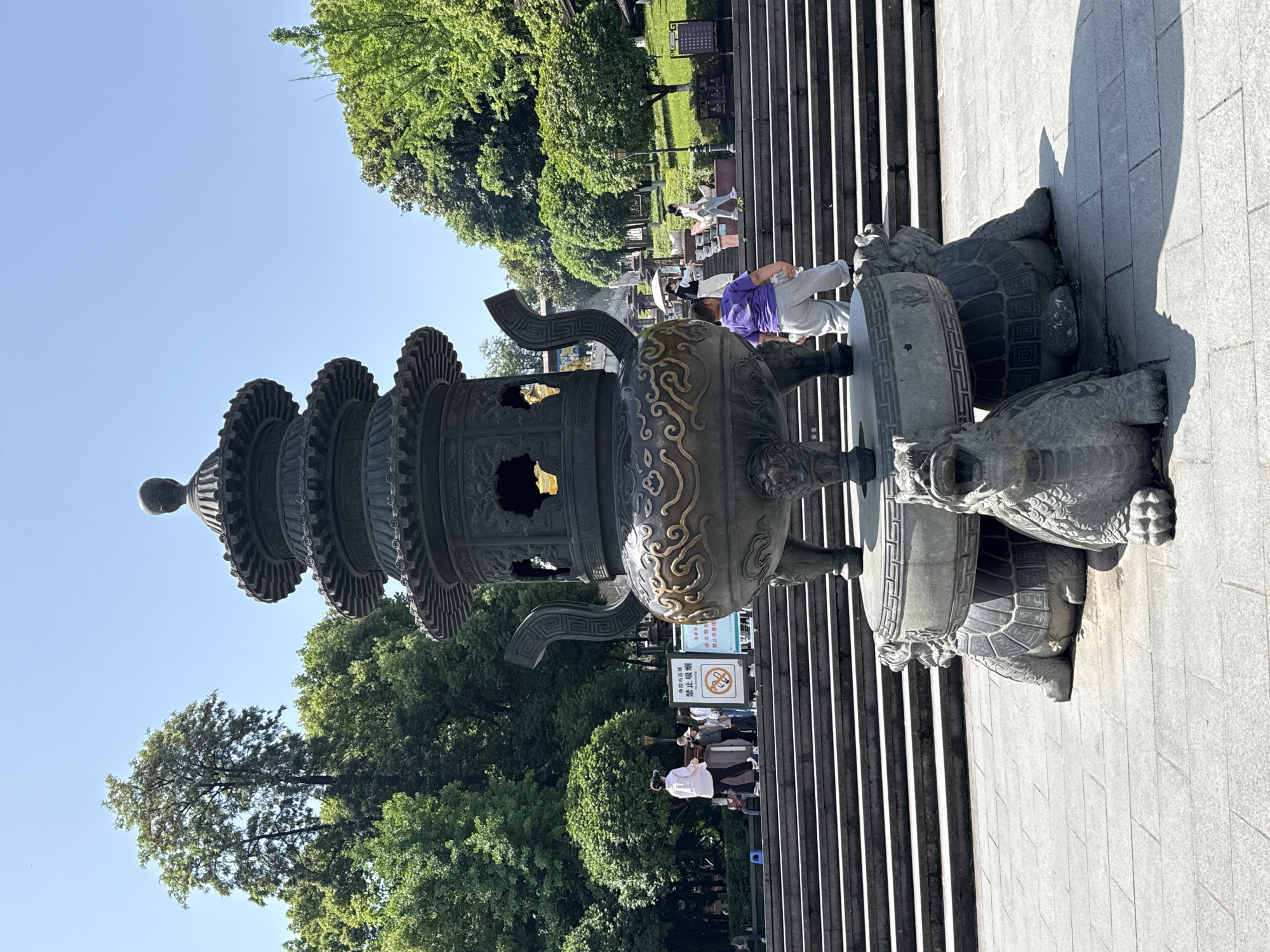 A bronze incense burner with a tiered top and ornate base is positioned in front of a set of stairs in Jiulongpo District, China. The burner is supported by sculptures of turtles. People are ascending the stairs, and trees are visible in the background.