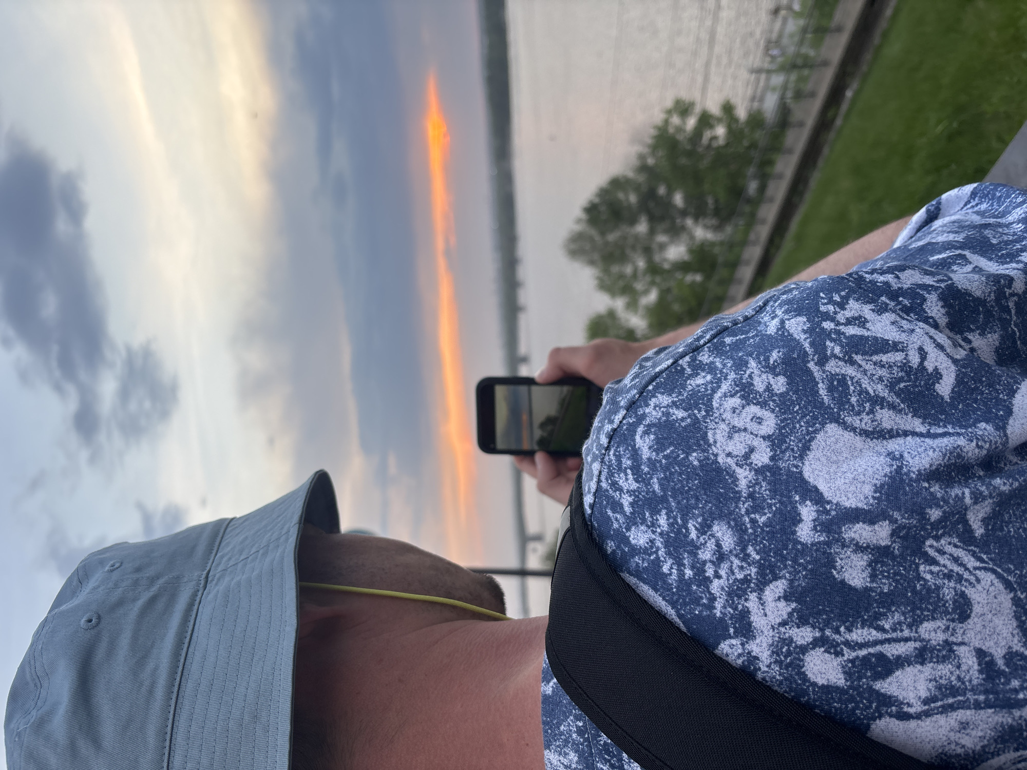 A person wearing a blue patterned shirt, a bucket hat, and a black strap is holding a cell phone, taking a picture of a sunset over what appears to be a body of water. The sky displays hues of orange, gray, and white. A green tree and a gray concrete embankment are visible to the right. The photograph is taken from a slightly elevated position, looking towards the horizon. Given the visual elements, including the wide river, trees, and embankment, the location is likely the Kama River embankment in the city of Perm, Russia.