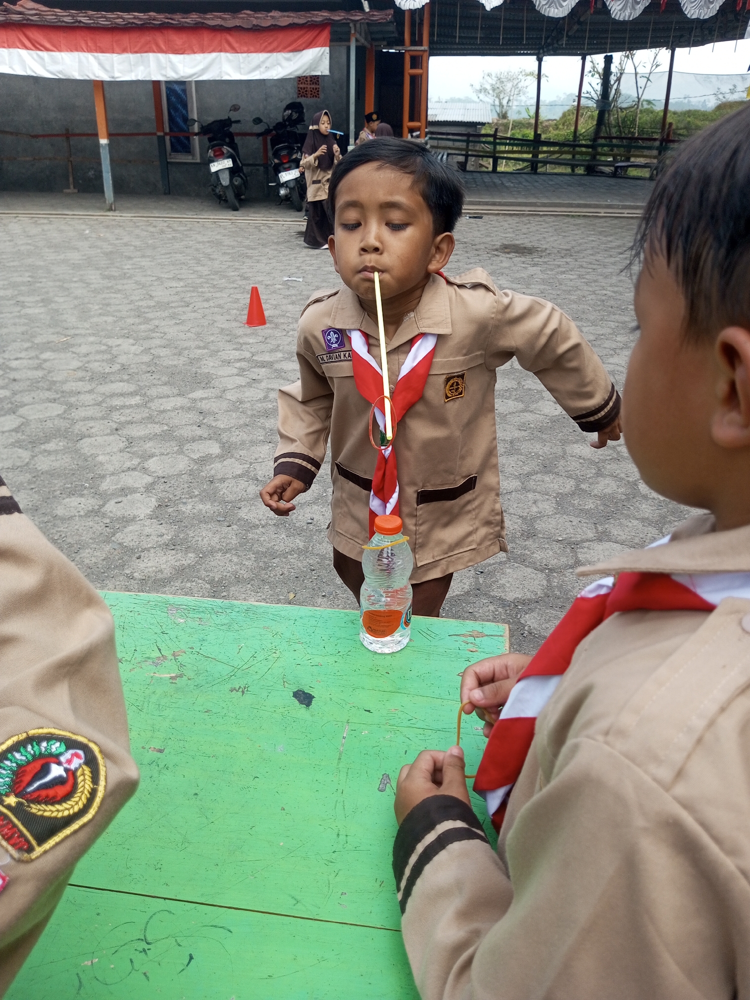 In Kalibeber, Indonesia, two young boys, dressed in tan scout uniforms with red and white scarves, are engaged in an activity. The boy in the foreground, facing away from the camera, is holding a yellow rubber band. The boy in the center, facing the camera, is holding a thin stick with his mouth and a plastic bottle of water is next to him on a green table. The environment appears to be an outdoor, paved area with a red and white banner overhead. In the background, two motorcycles are parked, and other children and adults are visible. The scene suggests a scout activity or game taking place outdoors.