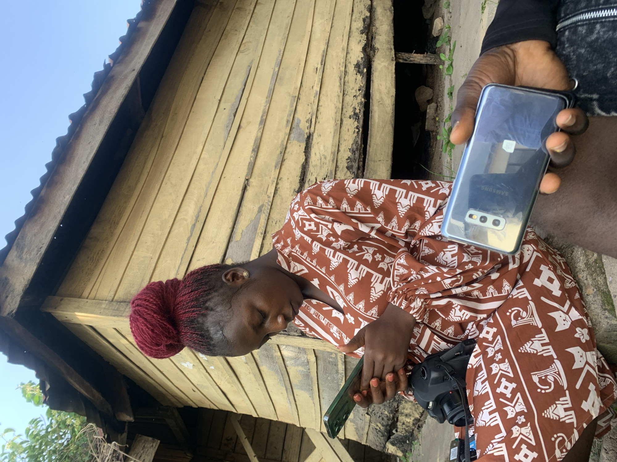 An outdoor, low-angle photograph captures two individuals in Jos, Nigeria, set against a weathered, light yellow wooden building with a dark corrugated metal roof and a clear blue sky. A young woman with dark skin and red braided hair, styled in a high bun, sits prominently in the left-center. She wears a brown and white patterned dress, featuring designs of cats and structures, and is engrossed in using a green-cased smartphone held in both hands. Her fingernails are painted orange, and she wears small, gold-colored flower stud earrings. A black Canon DSLR camera rests on her lap, alongside a smaller black electronic device. To the lower right, a dark-skinned arm and leg of a second person are visible, holding a silver Samsung smartphone that reflects the sky and a white rectangular object. The wooden planks of the building show signs of age, with peeling paint and visible gaps, while sparse green vegetation grows near the roofline and on the dirt ground beneath the structure.