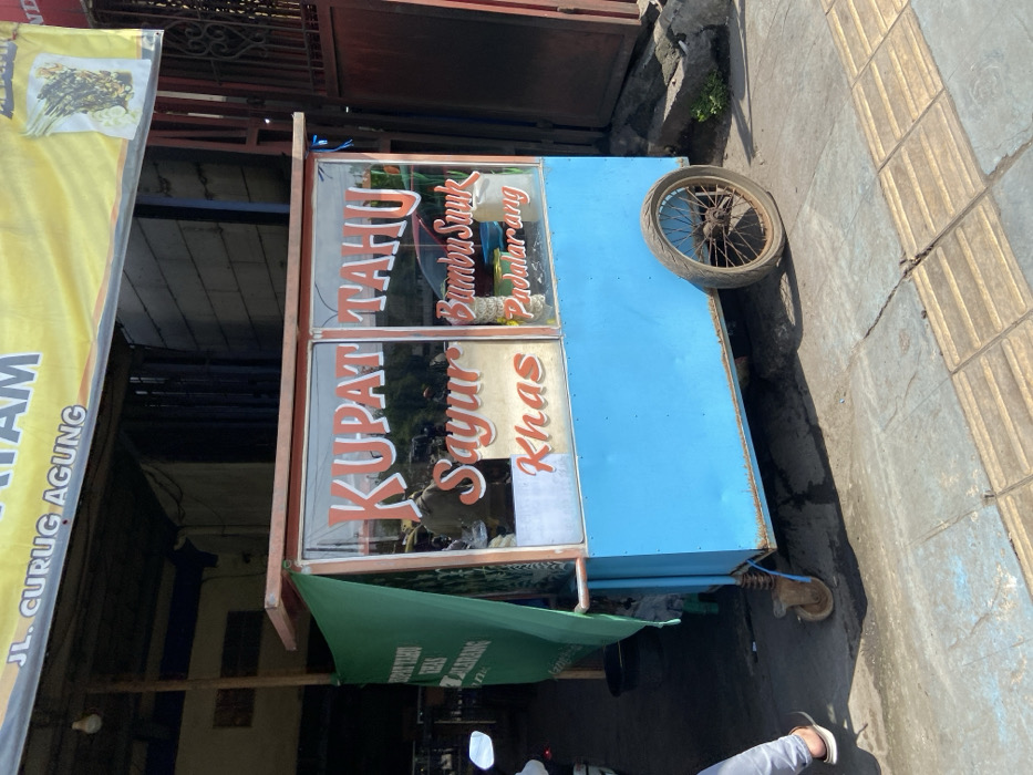 A food cart with a blue body and glass windows sits on the sidewalk in front of a building. The cart has a sign that reads "KUPAT TAHU Sayur Khas Bumbu Suuk Padalarang". The cart has a single, large wheel on the right side and a small wheel on the left. There is a green awning in the background. The cart is located on JL. Curug Agung in the city of Padalarang, Indonesia. The latitude and longitude coordinates are -6.845 and 107.498, respectively.