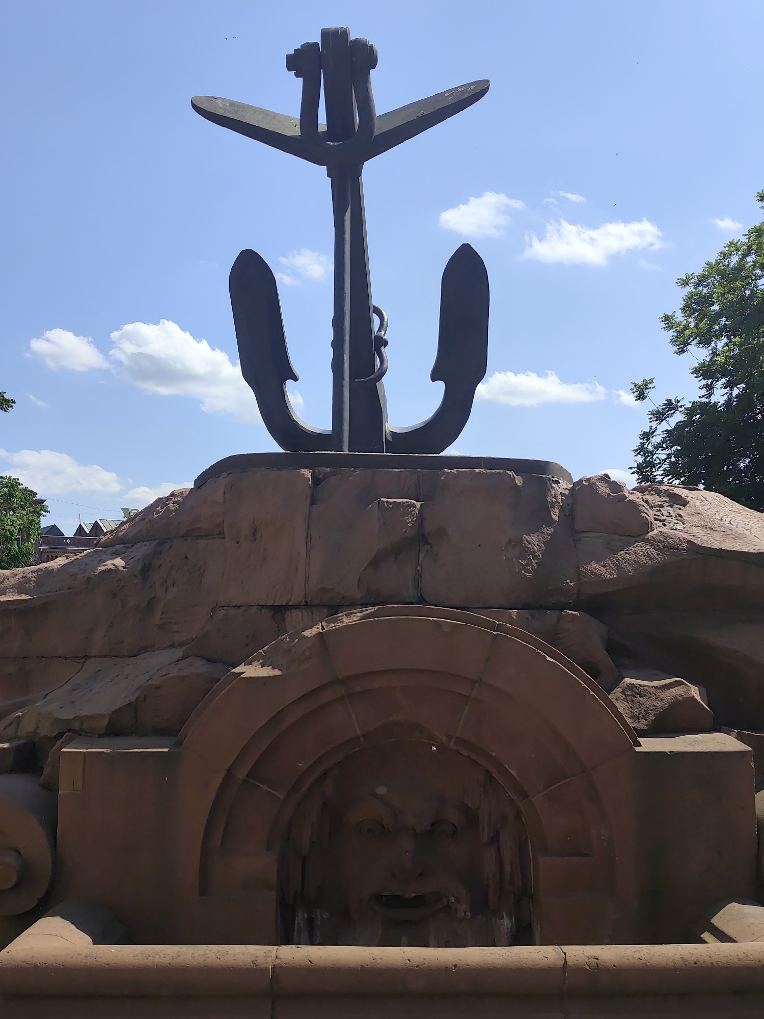 The image shows a large, dark metal anchor mounted on a reddish-brown stone structure. The anchor is positioned vertically, with its flukes spread outwards at the top. Below the anchor, the stone structure resembles a stylized rocky outcrop. An arched niche is carved into the lower part of the stone structure, containing a sculpted face that appears to be a grotesque or mythological figure with open mouth and a beard. The scene is set against a bright blue sky with scattered white clouds and a few distant birds. Green foliage from trees is visible on the right side of the image, and some buildings can be seen in the background to the left.
This monument is located in Szczecin, Poland, at the "Fountain of the Four Elements" on Plac Grunwaldzki. It is part of a larger fountain complex, with this particular section representing the element of water, symbolized by the anchor and the carved face, often interpreted as a sea god or Neptune. The fountain was designed by a Polish artist and is a prominent landmark in the city.