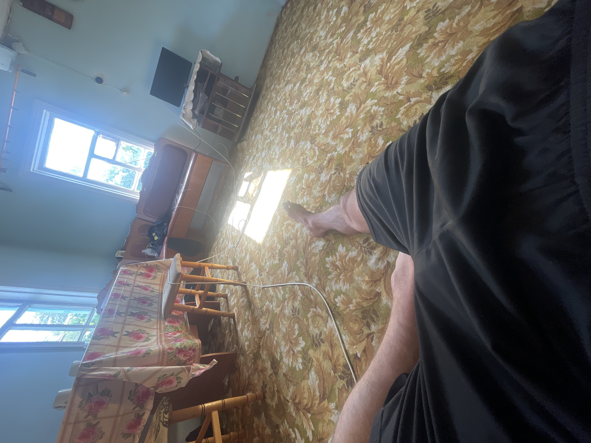 The image shows a first-person perspective of a room. In the foreground, black shorts and a person's legs are visible, positioned on a carpet with a floral pattern. The carpet features a dominant color scheme of green, yellow, and white. Sunlight streams in from a window on the right, illuminating a portion of the carpet.
In the background, a wooden dining table is draped with a tablecloth adorned with pink roses. Two wooden chairs are positioned at the table. Further back, a wooden bench with an orange cushion is visible, and a small black television sits on a wooden stand to the right. A window with multiple panes is present on the right side of the room, letting in bright daylight. The walls are painted a light blue. The environment appears to be an indoor living space, likely a dining or living room. The location is identified as Nikolayevskaya, Russia.