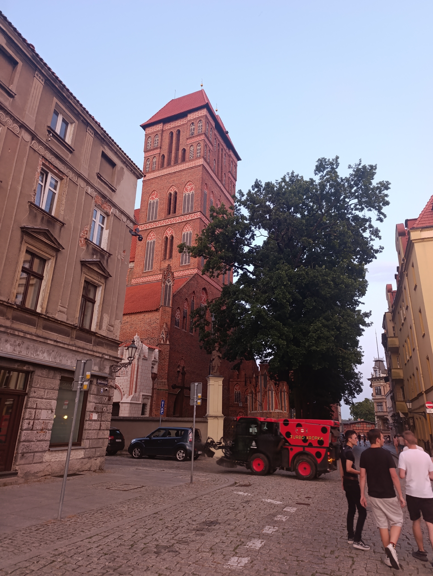 The image shows a street scene in Toruń, Poland, with a prominent brick tower of a cathedral in the background. The tower features Gothic architectural elements, including arched windows and decorative brickwork. In the foreground, a red street sweeping vehicle with ladybug-like polka dots and the text "TURBO KROPKA" is actively cleaning the cobblestone street. Several people are walking on the sidewalk and street. To the left, a multi-story building with windows and some signs is visible. To the right, another building with yellow facade stands. The sky is clear, suggesting it is late afternoon or early evening.