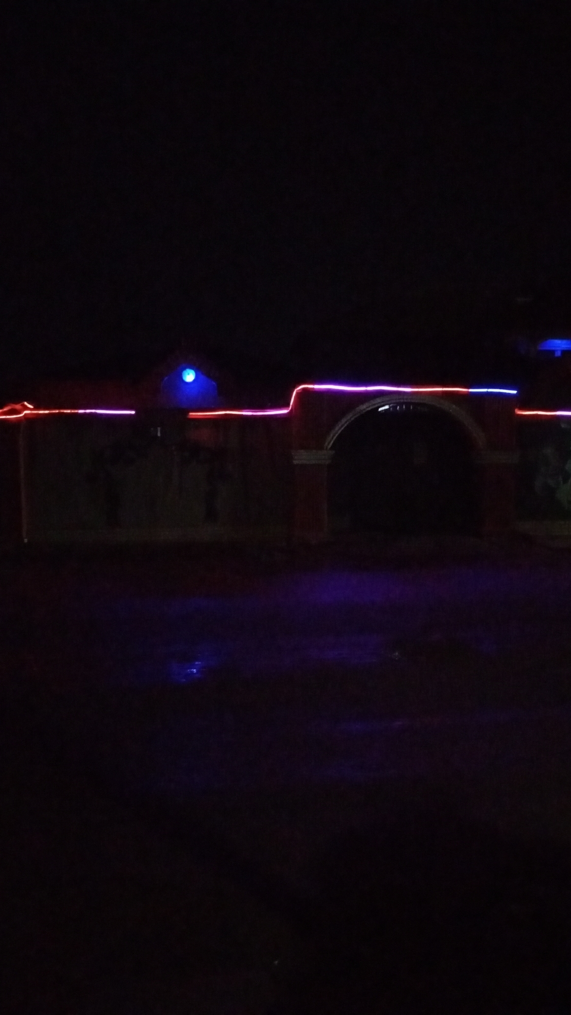 The image depicts a nighttime scene in Sabon Tasha, Nigeria. The main subject is a walled structure with an arched entrance, illuminated by red and blue string lights along its upper edges. There is a blue light atop the wall to the left. The foreground is dark, possibly a road or yard, with a faint purple hue. The environment is generally dark, suggesting limited ambient lighting. There are no visible people or other subjects. There are no specific actions or interactions apparent. The image focuses on the architectural structure and its decorative lighting within the urban landscape.