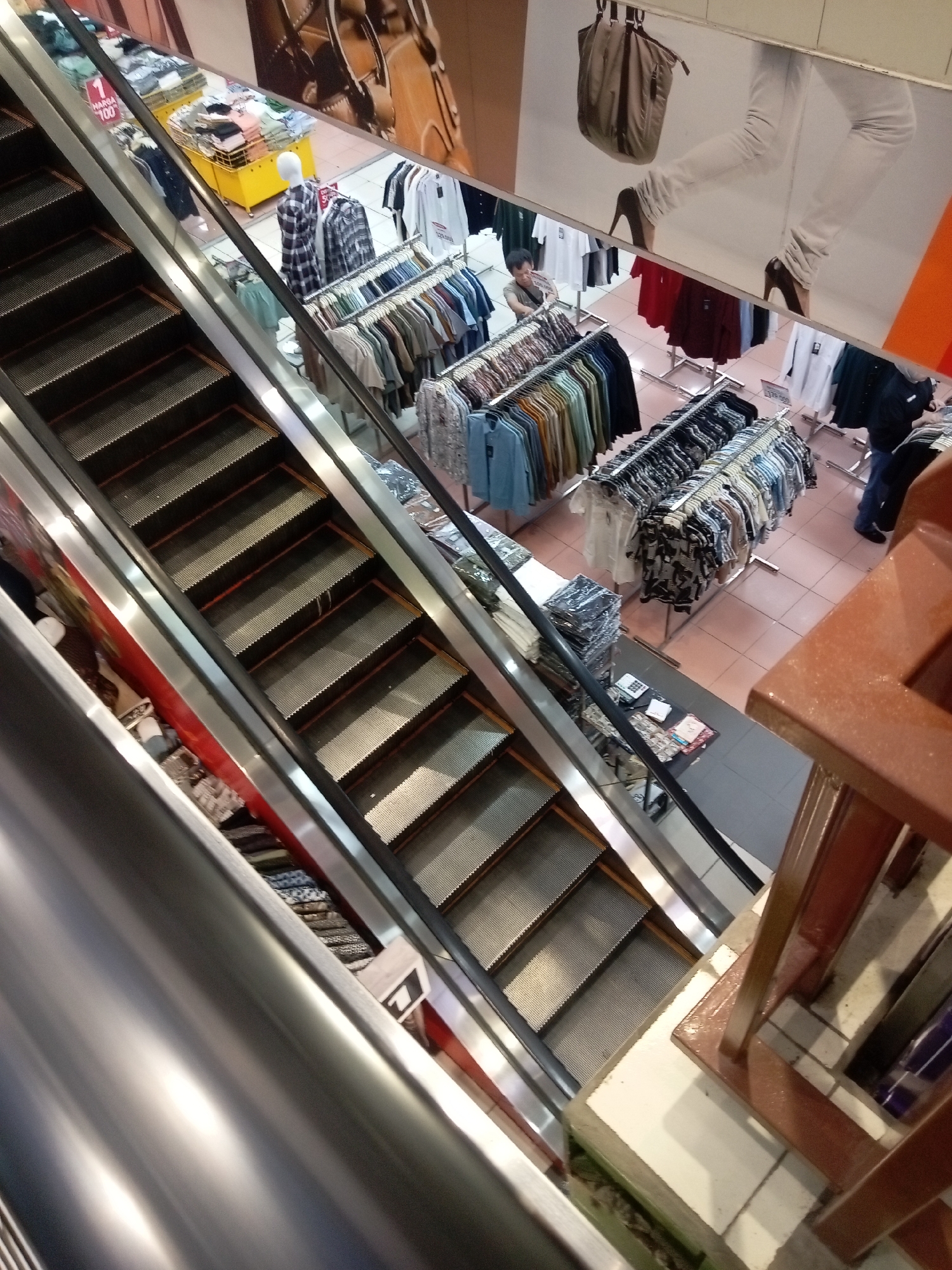 The image depicts an interior view of a retail establishment, likely a department store or clothing shop, in Batam, Indonesia. An escalator is prominently featured in the foreground, its metallic steps descending. Beyond the escalator, the scene unfolds into a retail floor with various clothing racks displaying a selection of apparel, primarily shirts and blouses in a variety of colors and patterns. A yellow display rack with folded clothing is visible, as is a mannequin dressed in a plaid shirt. In the background, a large advertisement featuring a woman's legs in white pants and high heels is displayed on the wall. Several individuals are present in the store, including a person looking at clothes on a rack and another person walking by. The flooring appears to be tiled. The overall impression is that of a busy commercial environment.