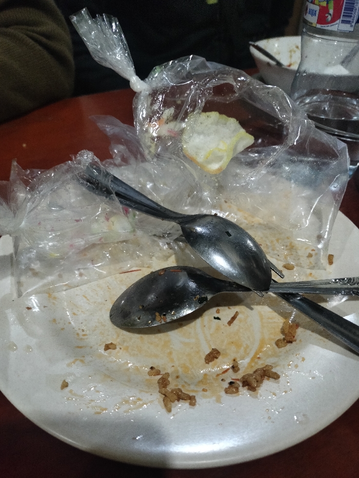 A close-up, low-light photograph captures the remnants of a meal on a dirty white plate. The plate is significantly stained with brownish-orange residue, likely from a sauce or gravy, and scattered small, irregular-shaped food pieces, possibly fried rice or similar grains. Two metallic spoons, also bearing visible food residue, rest on the plate.
Several clear plastic bags are prominent. One bag, tied at the top, contains a single piece of white, crinkled krupuk (cracker). Another clear plastic bag is partially visible underneath the spoons.
In the background, a dark brown table serves as the surface. A portion of a dark-colored garment, possibly belonging to a person, is visible on the upper left. On the right, a clear plastic water bottle is partially in view, with its label clearly displaying the brand name "AQUA" and a barcode, a common bottled water brand in Indonesia.
The image depicts a scene after eating, characterized by mess and food waste on the plate and spoons, along with plastic packaging. The lighting is dim, contributing to a somewhat subdued atmosphere. No active event is taking place, only the aftermath.