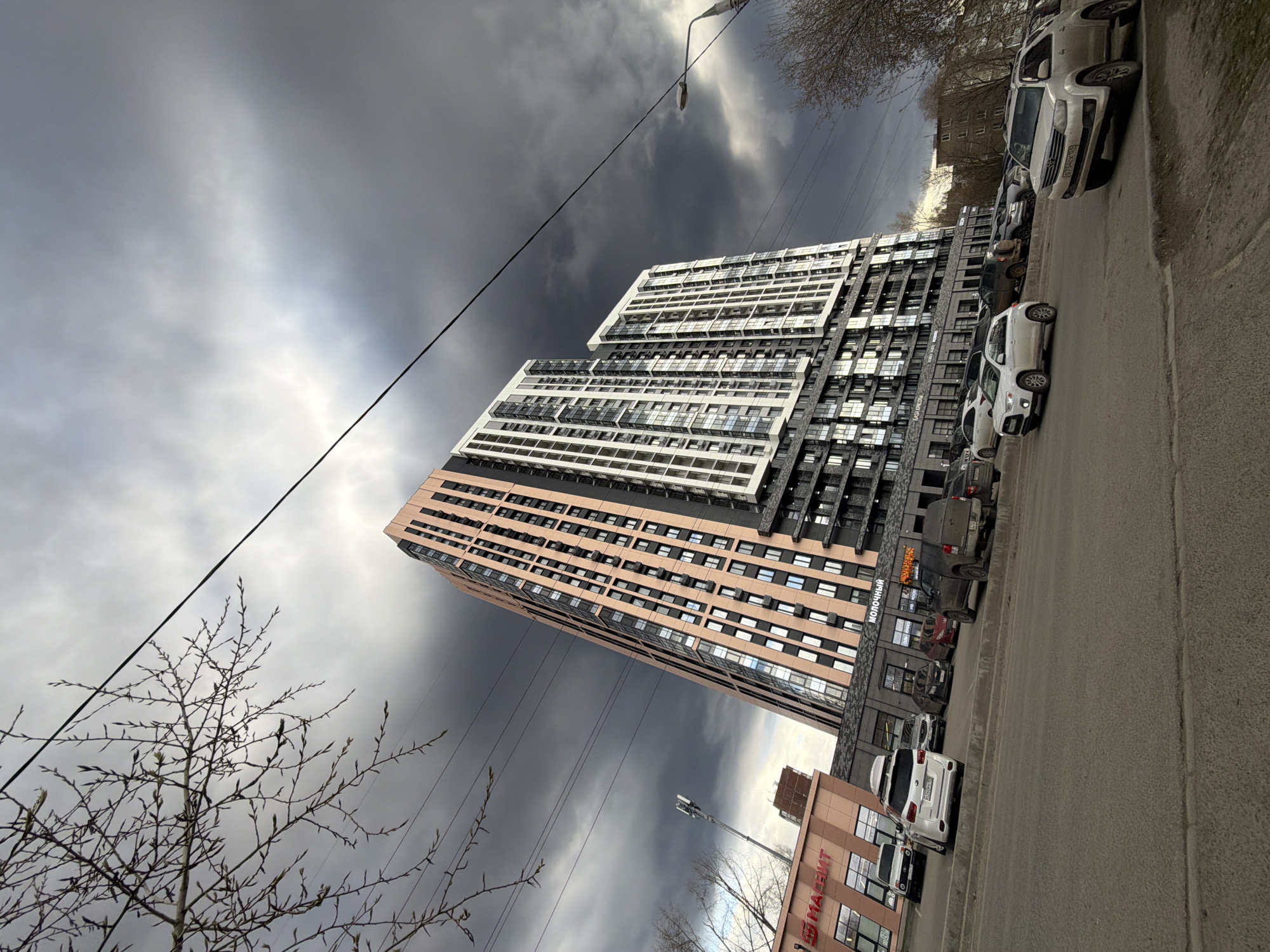 The image captures a street scene dominated by a tall, modern residential building under a dramatic, overcast sky. The photograph is taken from a low angle, looking upwards, and is vertically oriented (rotated clockwise).
The central subject is a multi-story building featuring a distinctive design with sections in light white/grey, dark black, and a reddish-brown/tan color. It has numerous windows and balconies. To the right of the building, a paved street runs parallel to it, with several cars parked along the curb. These vehicles include various models, primarily SUVs and sedans, in colors like white, grey, and black. A white SUV is prominently featured in the upper right section of the image.
The sky is heavily clouded, presenting a mix of dark grey, almost black, storm clouds contrasting with lighter, brighter patches. A street light or utility pole with overhead wires stretches diagonally across the frame. In the foreground on the left, a bare tree with small buds suggests either late winter or early spring. On the far right, a smaller, lower-rise building with a reddish-brown facade and large windows is partially visible.
Visible text on the side of the main residential building near its base reads "МОЛОЧНЫЙ" (MOLOCHNЫY), likely indicating a commercial establishment or the name of the complex. On the smaller building in the foreground, the text "МАГНАТ" (MAGNAT) is visible. A license plate on one of the white SUVs shows the region code "159", consistent with Perm Krai, Russia. The scene depicts an urban environment, likely a street in Perm, Russia, during daytime under heavily overcast conditions. There is no specific activity or event taking place; it's a static view of city infrastructure and weather.