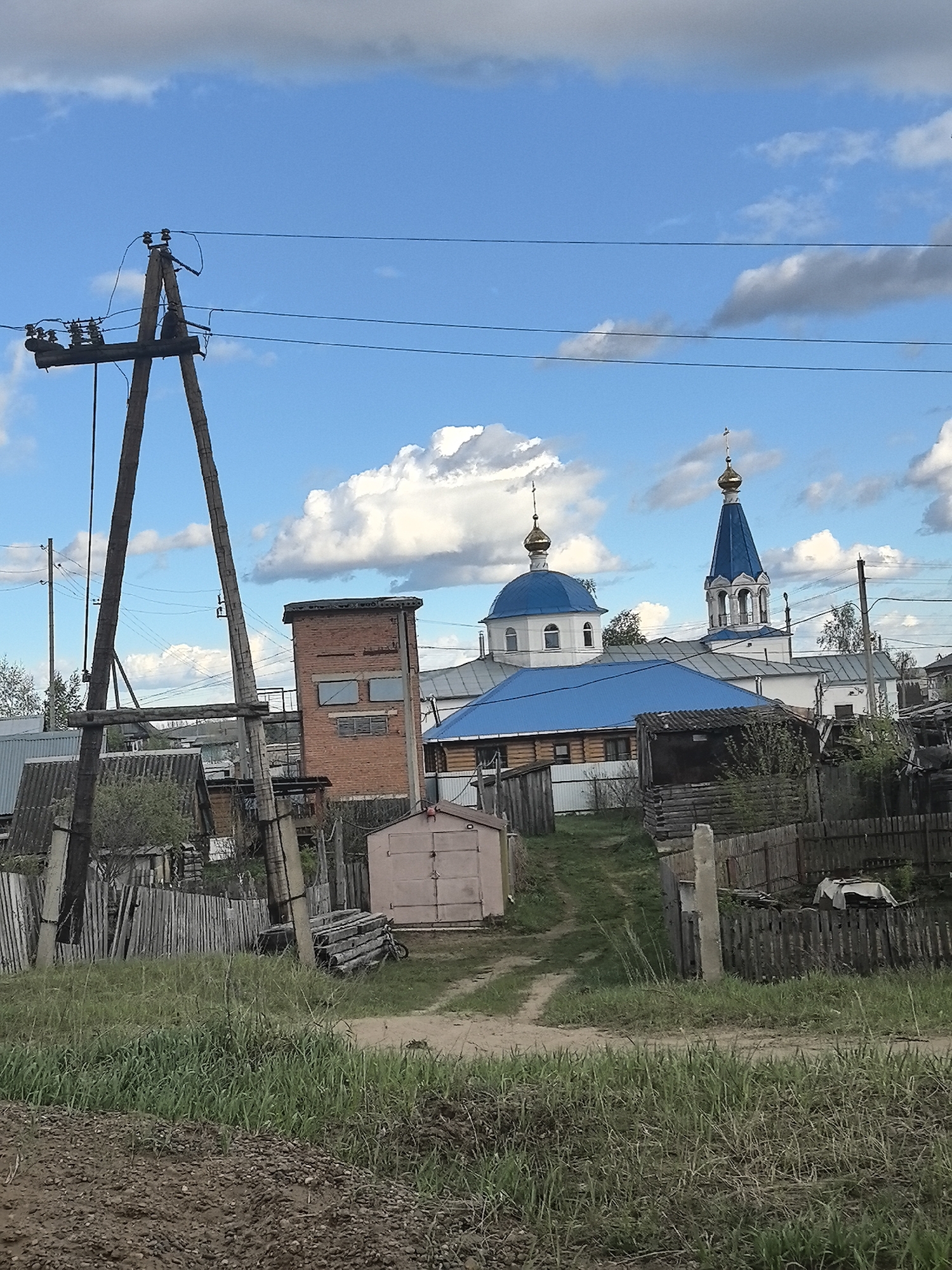 The image depicts a rural scene in Noviy, Russia, under a partly cloudy sky. An Orthodox church with blue domes and a bell tower stands in the background. In the foreground, there are wooden fences, a brick building with a few windows, a small shed, and overgrown grassy areas alongside a dirt path. An old wooden utility pole is visible on the left side of the image. A bicycle is lying on the ground near the small shed. The scene conveys a sense of simplicity and rural life.