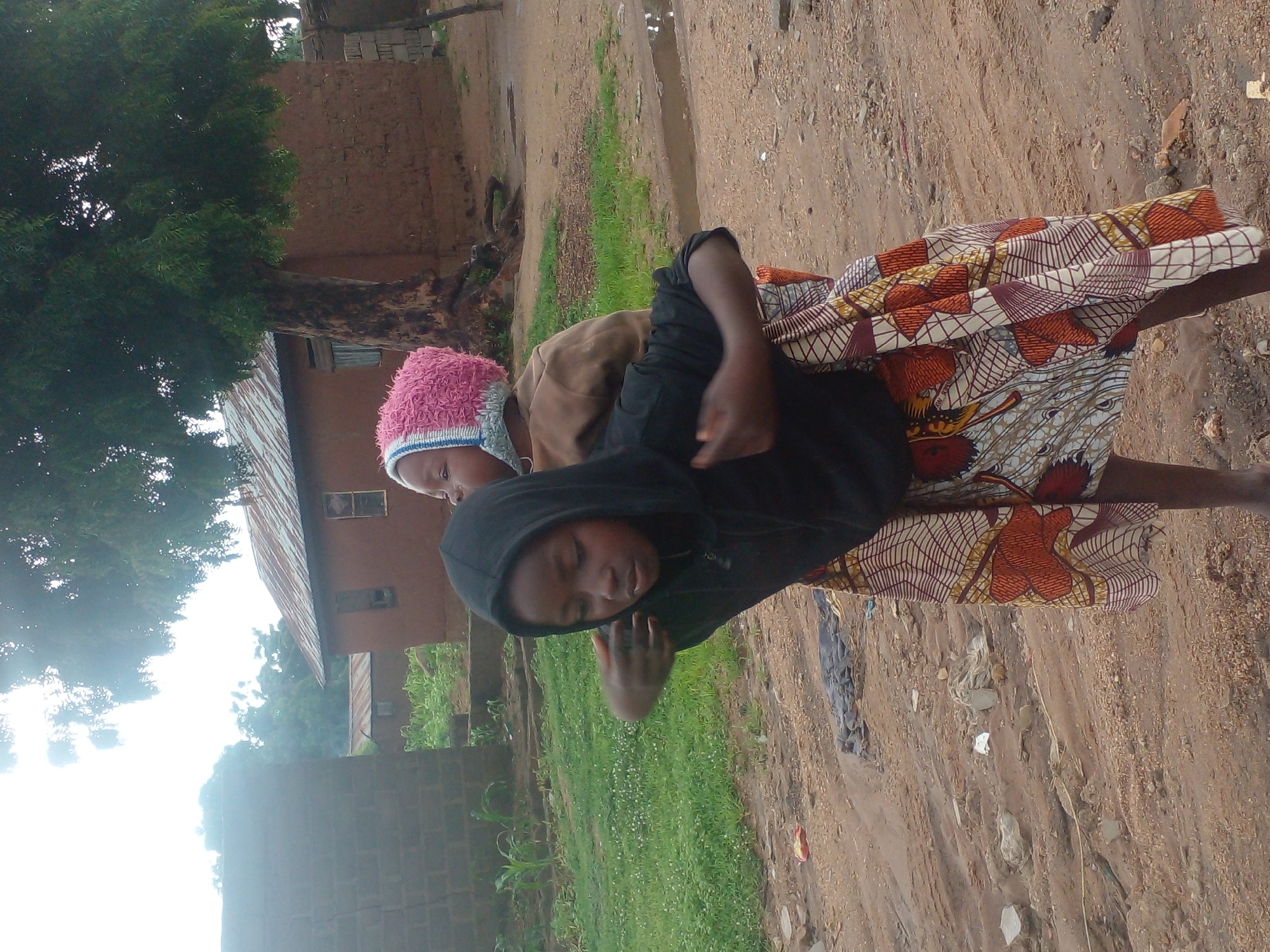 In Langtang, Nigeria, two young girls stand on a dirt path. The girl in front, wearing a black hooded sweatshirt and a colorful patterned skirt, has her arm around the shoulders of the girl behind her. The second girl, wearing a brown shirt and a pink knitted hat with a blue band, is partially obscured. They are positioned in front of a tree and several rustic, mud-brick buildings with corrugated metal roofs. The ground is a mixture of dirt, small stones, and some scattered debris, with a puddle of water visible to the right. The overall environment suggests a rural or semi-urban setting.