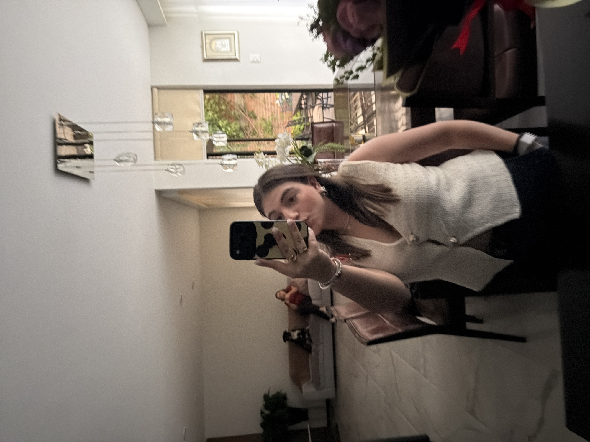 A young woman is taking a selfie in an indoor environment that appears to be a room with an open design. The location is Barrio de Nuevo León, Mexico. The room has white walls, with a piece of artwork that consists of triangular shapes. A chandelier with multiple light fixtures is suspended from the ceiling. A window or opening shows a view of greenery, possibly trees, and sunlight. There is a picture frame on the wall. A bouquet of flowers sits on a piece of furniture. The woman is wearing a light-colored top and dark bottoms. She is holding a cell phone with a distinctive case. Furniture and decorative items are present, including a plant and decorative figures on a shelf or table. The overall ambiance is casual and the environment seems well-lit.