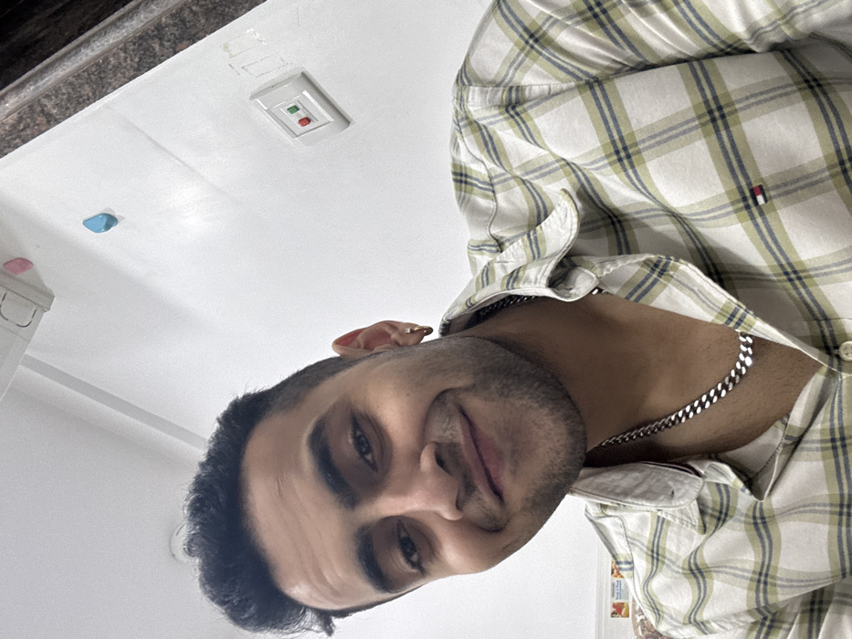 A young adult male is depicted in a close-up, upward-angled selfie or portrait, set within an indoor environment. He has dark hair, a stubble beard, and is looking directly at the viewer with a slight smile. He wears a cream-colored collared shirt with a green and blue plaid pattern, featuring a small "Tommy Hilfiger" flag logo on the left chest. A thick, silver-colored Cuban link chain necklace is visible around his neck, and he has a small gold-colored hoop earring in his right ear.
The background consists primarily of a plain white wall. On the upper right, a white electrical switchboard with one green and one red push-button switch is mounted. Above this, the wall shows some light surface wear or marks. Higher and slightly to the left, a small, light blue, roughly teardrop-shaped object is affixed to the wall. Near the top edge of the frame, a partially visible white and pink object, possibly a light fixture or appliance, is mounted near the ceiling. To the far right, a dark brown, textured vertical element, likely a door frame or structural pillar, is visible. On the far left, a sliver of a light-colored, possibly tiled or patterned wall surface is present at the bottom. The interior architectural elements and electrical fittings are consistent with residential or commercial spaces found in India. While the image does not provide specific landmarks, the context of the interior environment and the individual's appearance are consistent with a setting within the city of Kolkata, India, though a precise location cannot be identified.