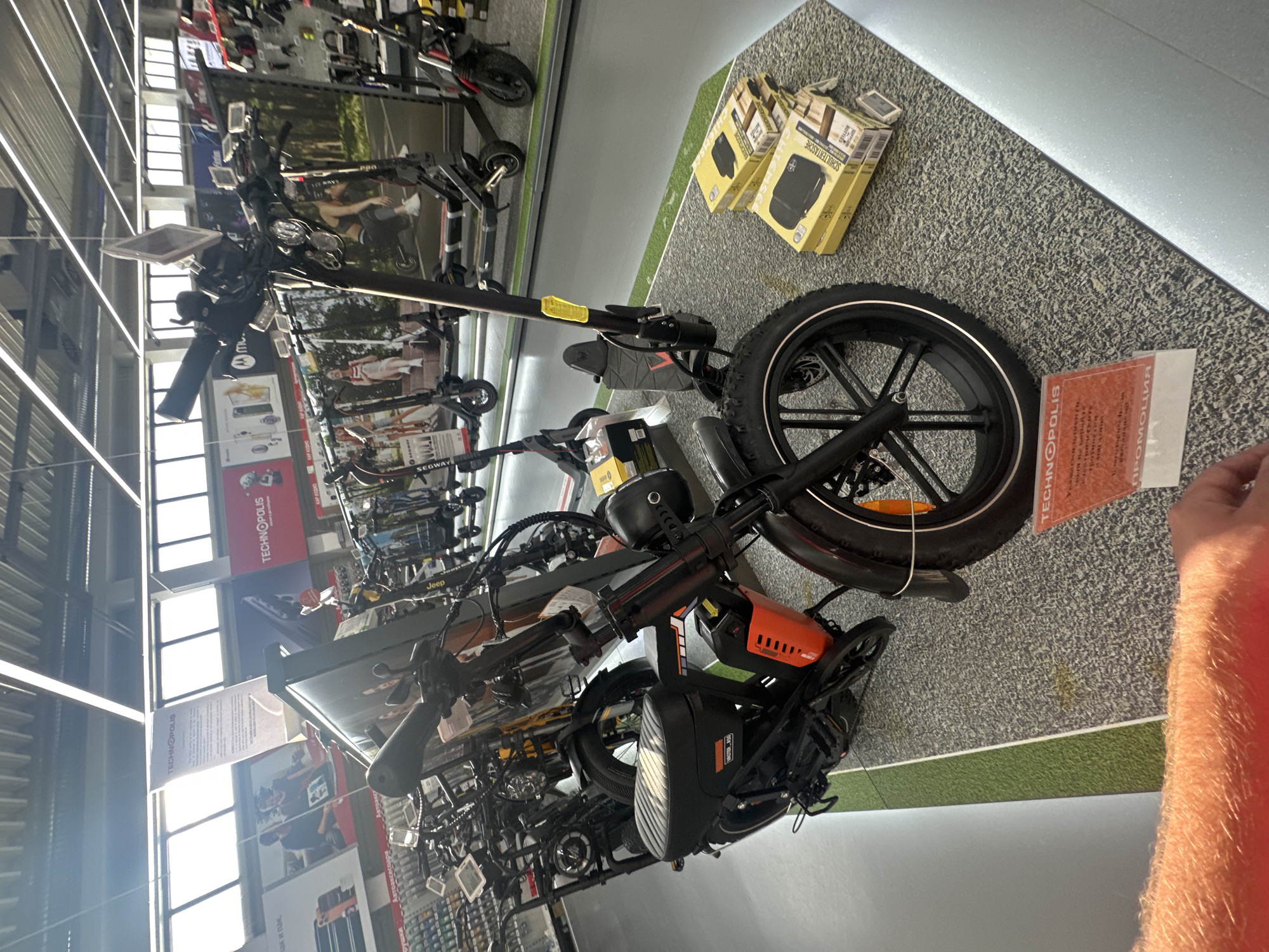 The image shows an indoor retail store, likely a bicycle or electronics store, in Sofia, Bulgaria. The primary subject is a black electric scooter with orange accents, displayed prominently on a textured floor. The scooter has a large, rugged tire and a visible battery pack. Several other scooters and bicycles are visible in the background, suggesting a wide selection of products.
In the upper right corner of the image, there are three yellow boxes stacked on top of each other. One box has text printed on it, which appears to be in German, with the words "SCHULTERTASCHE" visible, indicating it's a shoulder bag.
The store features bright overhead lighting, typical of a retail environment. Large windows are visible on the left side of the image, allowing natural light to enter, suggesting it is daytime. Posters and signs are hung throughout the store, including some in what appears to be Cyrillic script. One prominent red sign reads "TECHNOPOLLIS". Another white banner displays text in both Cyrillic and Latin alphabets.
A person's arm, covered in fair skin and light hair, is visible on the right side of the frame, reaching towards the scooter or a sign. The arm is wearing a bright orange sleeve or shirt.
The overall scene depicts a commercial setting where electric scooters and related items are being sold. The presence of multiple products and the retail environment suggest a shopping experience is taking place or available. The time of day is clearly daytime, and the setting is indoors.
