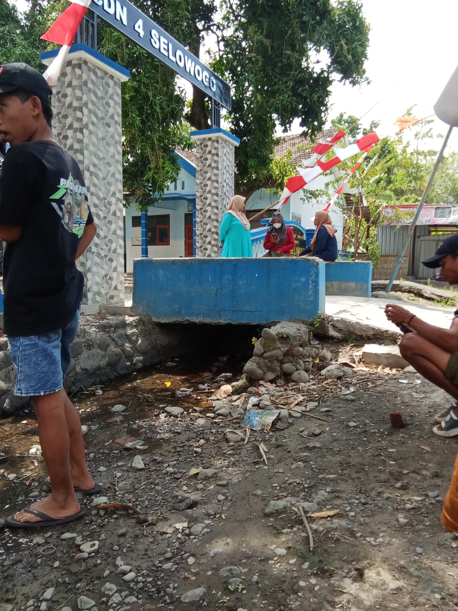 The image depicts the entrance of SDN 4 Selowogo, a school located in Trebungan, Indonesia. In the foreground, a young person wearing a black t-shirt and denim shorts stands on the left, facing away from the camera. Their feet are clad in flip-flops. To the right, another individual is crouched down, holding a mobile phone.
Behind the foreground figures, a low, blue concrete structure runs across the mid-ground, with a small stream flowing beneath it, littered with rocks and debris. In the background, three individuals are seated on the blue structure. One person on the far left wears a bright teal hijab and turquoise dress. Next to them, a person in a red headscarf and mask looks at their phone. To their right, another individual in a brown hijab and dark blue top is also seated.
The school building is visible behind the seated individuals, with a light blue facade and red-framed windows. The entrance is marked by a stone pillar and a black sign displaying "SDN 4 SELOWOGO" in white lettering. Red and white Indonesian flags are attached to poles near the entrance. The environment is outdoors, with trees providing shade and a bright, overcast sky. The ground is a mixture of dirt, rocks, and some scattered litter.
