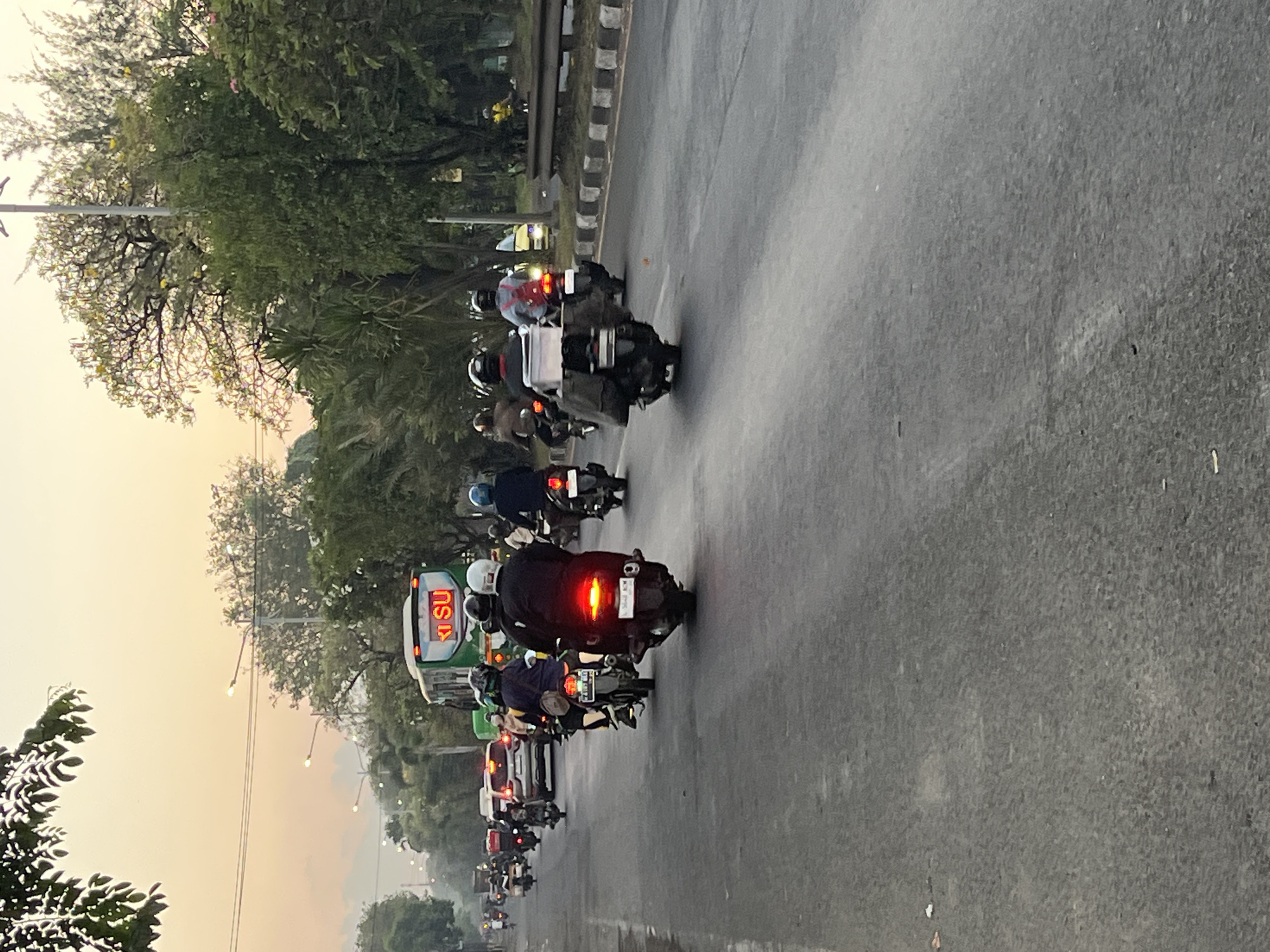 The image depicts a street scene in the city of Ngaban, Indonesia, during what appears to be the late afternoon or early evening, judging by the warm, muted light and the soft hues in the sky. The primary focus is a busy road with numerous motorcycles and several cars. The asphalt road is wet, indicating recent rain.
In the foreground, several motorcycles are visible, moving away from the viewer. The rider of the closest motorcycle is wearing a dark jacket and a white helmet. The motorcycle has a visible license plate that reads "BD 5648 XEM". Behind this motorcycle, another is slightly to the left, also with a rider wearing a dark jacket and a dark helmet. Further back, more motorcycles are in motion, some with passengers. Red taillights are illuminated on several of these vehicles, suggesting movement and braking.
A large, green bus with "K'SU" written in illuminated red letters on its front is a prominent element in the mid-ground, on the left side of the road. The bus appears to be in motion. Cars are also present, including a silver vehicle in the left lane, with its brake lights illuminated.
The environment is characterized by a line of trees and vegetation lining the right side of the road. Streetlights are visible along the road, some of which are illuminated. The background features more trees and a hazy sky with soft, diffused light. The overall impression is one of a typical urban commute in Indonesia, with a high volume of motorcycle traffic.