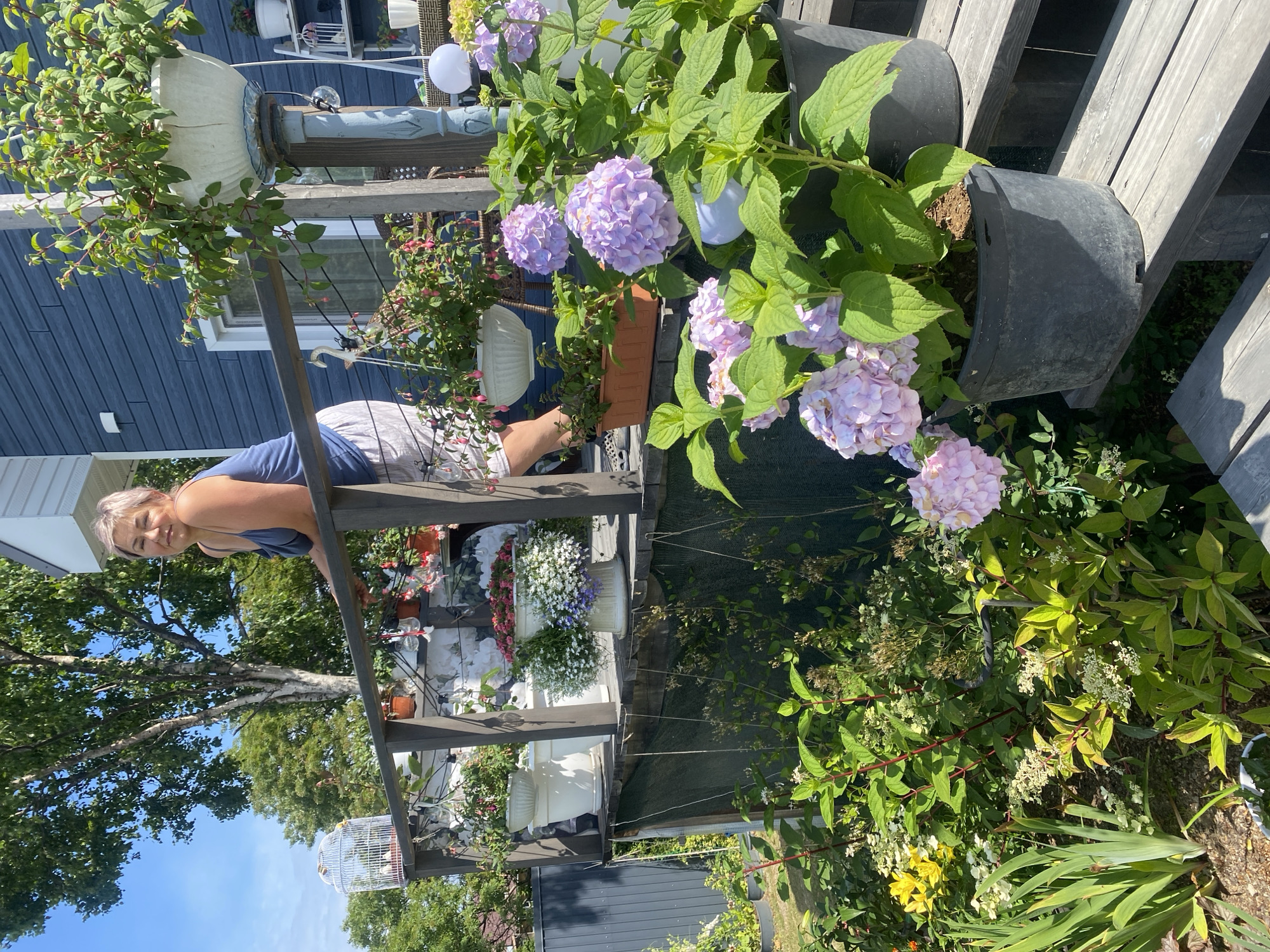 A middle-aged woman with blonde hair stands on a wooden deck, leaning on a railing. She wears a blue tank top and patterned shorts. The deck is decorated with numerous potted plants, including large hydrangeas in bloom, and other flowering plants. A blue-sided house is visible behind her. The scene is set outdoors on a sunny day, with lush greenery and trees surrounding the area. The woman appears to be in Tret'ya Pad', Russia, based on the provided context.