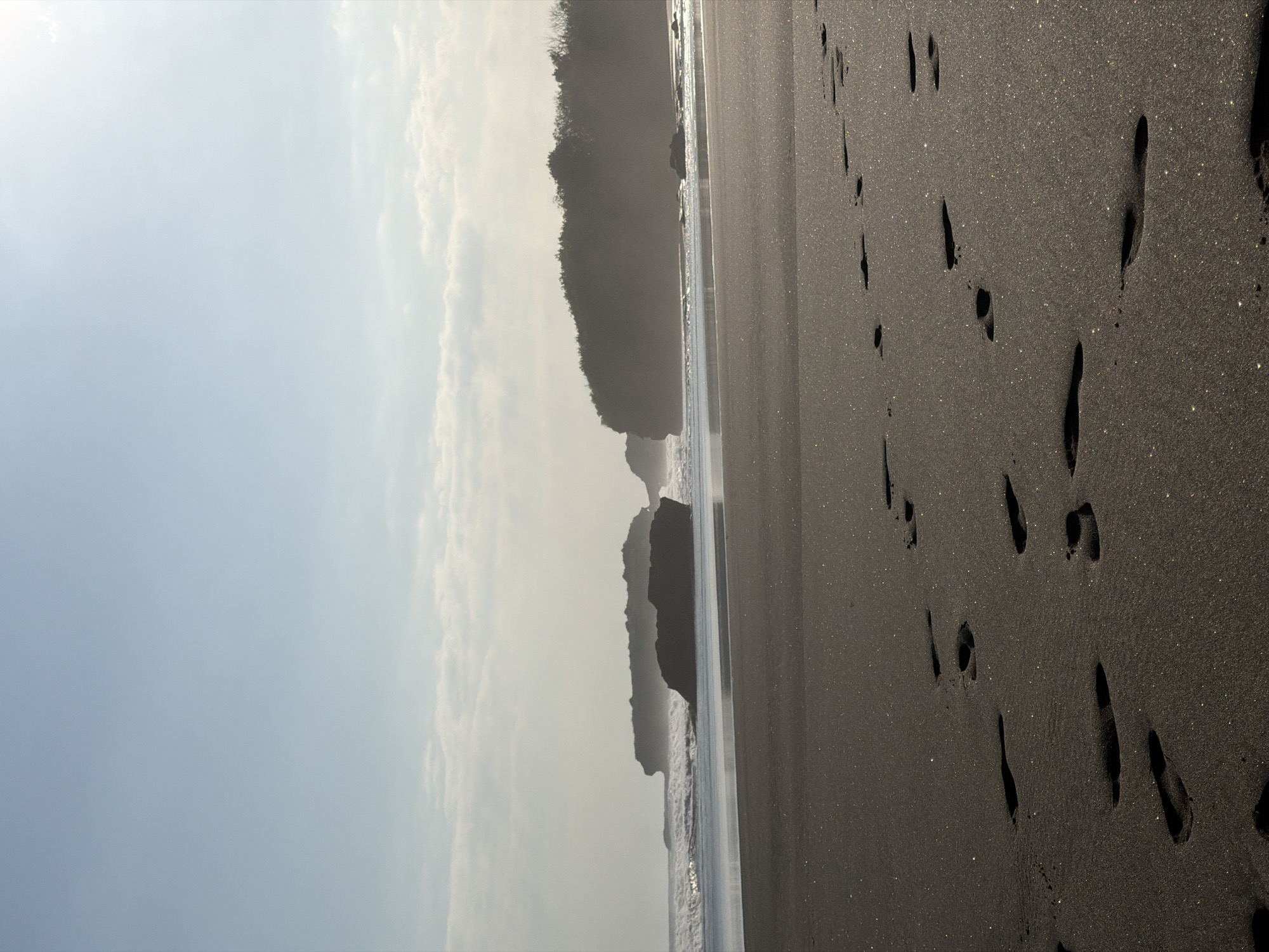 The image depicts a coastal landscape in Selemadeg Timur, Indonesia, under a bright sky. The upper two-thirds of the frame are dominated by a clear, light blue sky with faint, scattered white clouds, particularly near the horizon and in the upper right quadrant where the sun's glare is visible. The midground features a dark ocean with breaking waves, notably around several large, dark rock formations or small islands situated offshore. To the right, a darker, tree-covered landmass or cliff line extends, partially obscured by a subtle atmospheric haze. The foreground consists of a wide expanse of dark, possibly volcanic, sand. Numerous human footprints are clearly impressed into the sand, extending from the bottom left towards the right, indicating recent passage. The sand exhibits a distinct sparkling or glittering effect where it catches the sunlight, suggesting the presence of reflective mineral particles. No individuals are visible in the scene.