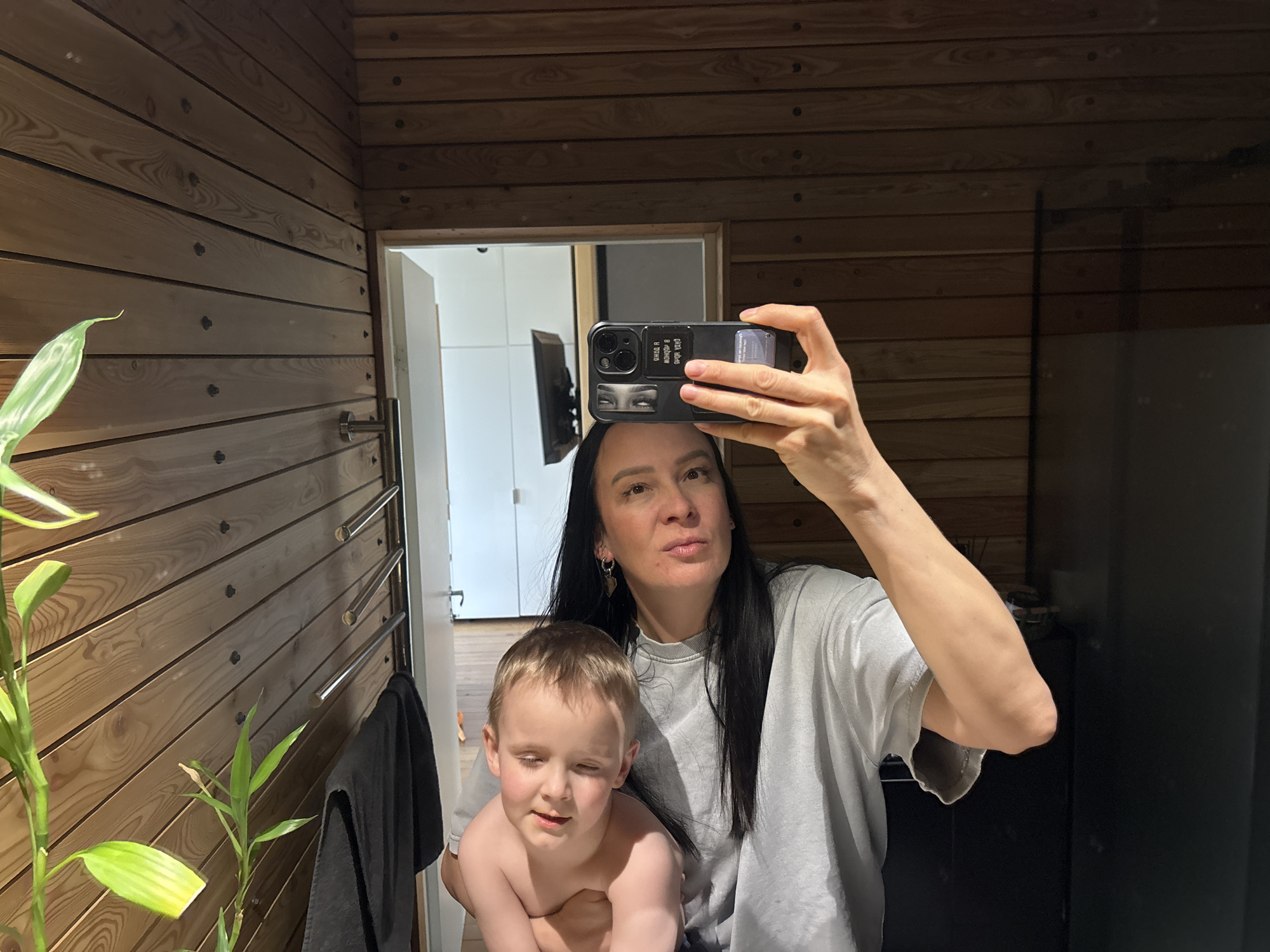 A woman and child take a selfie in a bathroom. The woman is fair-skinned with long black hair, wearing a gray sweatshirt. She holds a black smartphone up to take the photo. The child, a young boy with blond hair, is shirtless and winking. The bathroom has walls of horizontal wooden planks. A green plant is visible on the left, along with a towel rack and dark towel. A doorway leading to another room with white cabinets is visible in the background. The image was likely taken in a bathroom in Chaikovsky, Russia, given the prompt's request.