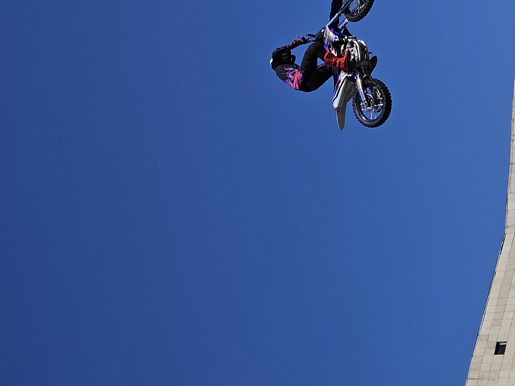 A motocross rider in mid-air is captured against a clear blue sky, with the upper portion of a light-colored building visible at the bottom of the frame. The rider is wearing a black helmet, a black and pink flame-patterned jersey, black pants, and bright red boots. They are on a dirt bike with purple and silver accents, performing a stunt with the bike tilted. The building appears to be made of rectangular blocks and has a small dark window. The image is taken from a low angle, looking up at the rider and the sky. The provided information states the location is Ulyanovsk, Russia, but this specific detail cannot be visually confirmed from the image content alone.