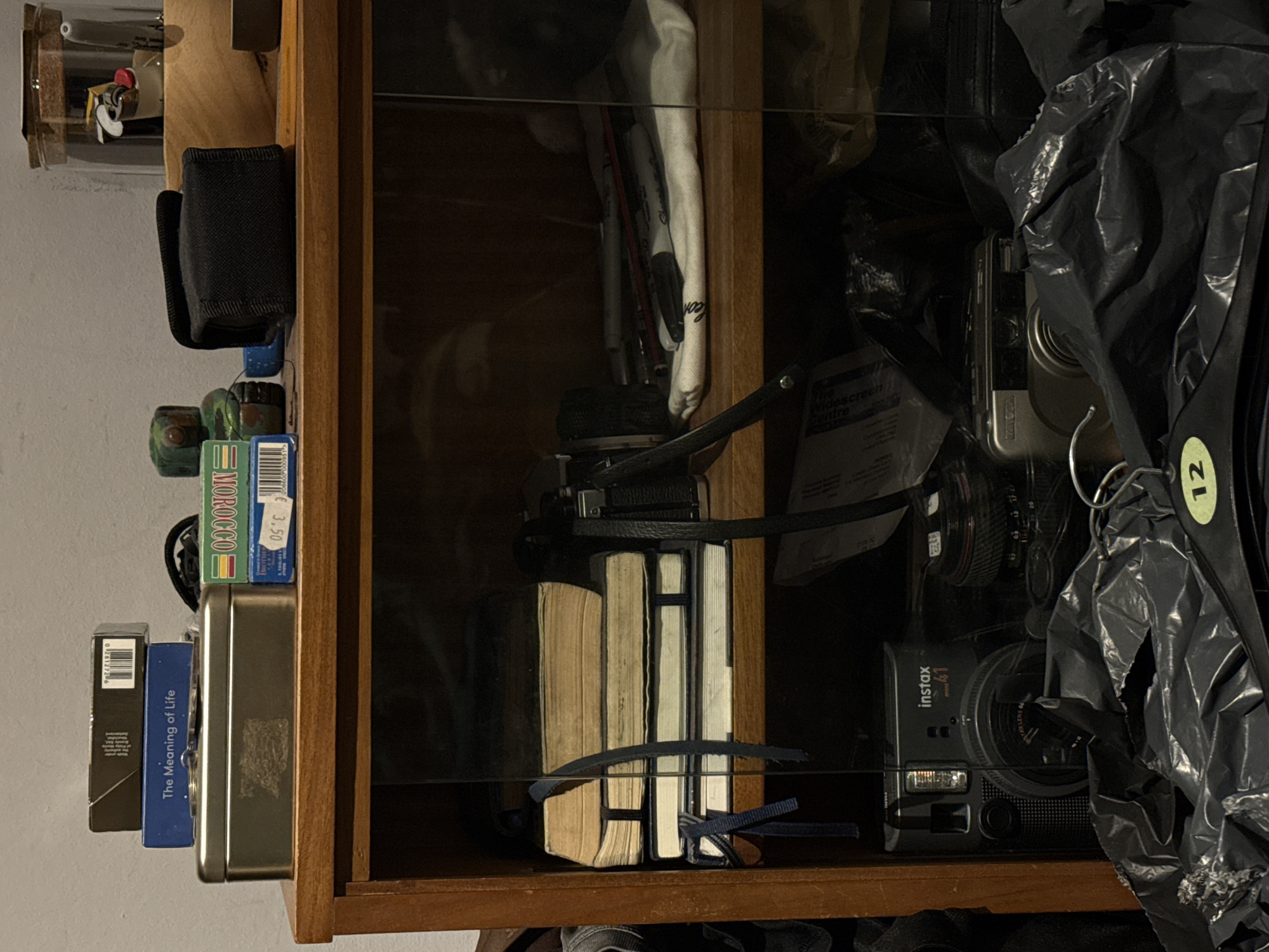 The image shows the contents of a cluttered wooden cabinet, likely indoors. On the left, various small items are stacked on a shelf, including a black pouch, small boxes labeled "MOROCCO" and "The Meaning of Life," and a silver-colored tin. Inside the cabinet, visible through a glass pane, are several books bundled together with a blue strap, a vintage camera with a leather strap, a Minolta camera, and a newer "instax mini 41" camera. There are also pens and a white cloth. A black plastic bag partially obscures the right side of the cabinet. A clothing hanger with a tag marked "12" hangs within the bag. A piece of paper is partially visible, with text related to "The Widescreen Centre" and "Leon."