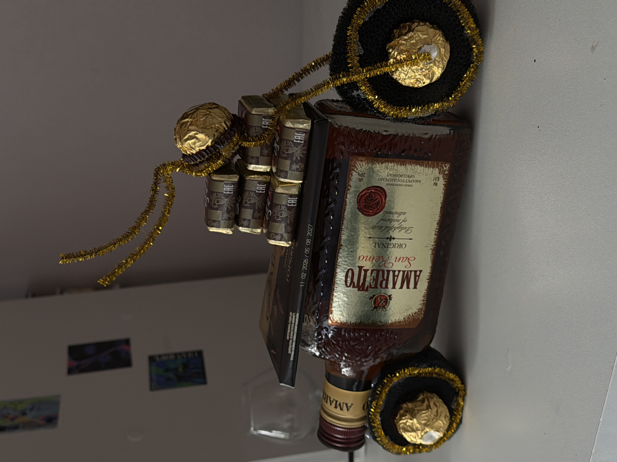 Here is a detailed description of the image:
**Subjects and Objects:**
* **Bottle of Amaretto San Remo liqueur:** The bottle is brown and labeled "Amaretto San Remo Original." There are labels in both English and Russian. The Russian label indicates that it is "AMAPETTO CAH PEMO ORIJINAL."
* **Ferrero Rocher chocolates:** Two of the chocolates, still in their gold wrappers, are positioned at the top and bottom of the bottle.
* **Candies:** Wrapped candies stacked on the top and side of the bottle.
* **Gold Tinsel:** Gold tinsel is wrapped around the candies and the bottle.
* **Refrigerator:** The bottle is near a refrigerator, as seen in the background.
* **Artwork:** Small pieces of artwork are adhered to the side of the refrigerator.
**Environment:**
* **Indoor setting:** The photograph appears to be taken indoors, possibly in a kitchen or living area, given the presence of a refrigerator.
* **Background:** The background is slightly blurry.
**General Description:**
The image is a close-up shot of an Amaretto San Remo bottle decorated with Ferrero Rocher chocolates, candies, and gold tinsel, suggesting it may be a gift or a decorative item. The bottle is placed near a refrigerator. The presence of Russian text on the label could suggest the image was taken in a Russian-speaking country.