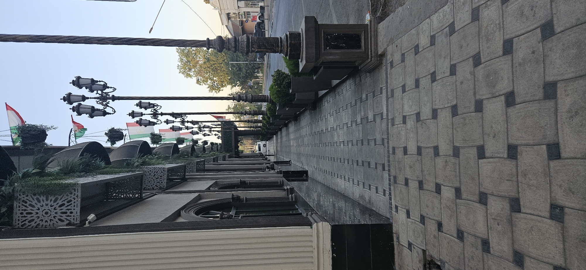 This is a low-angle, full outdoor shot of a sidewalk in Dushanbe, Tajikistan. The sidewalk is paved with decorative grey and black bricks laid in a basketweave pattern. To the left of the sidewalk, the facade of a building with classical columns and arched doorways is visible. Along the facade, there are decorative planters with hanging greenery and several flags, possibly representing Italy and Tajikistan, are displayed. The sidewalk is lined with ornate street lamps and planters filled with green shrubs. In the background, a street with cars and buildings is visible, along with trees and a clear sky. The scene appears to be in a city setting, likely a commercial or public area.