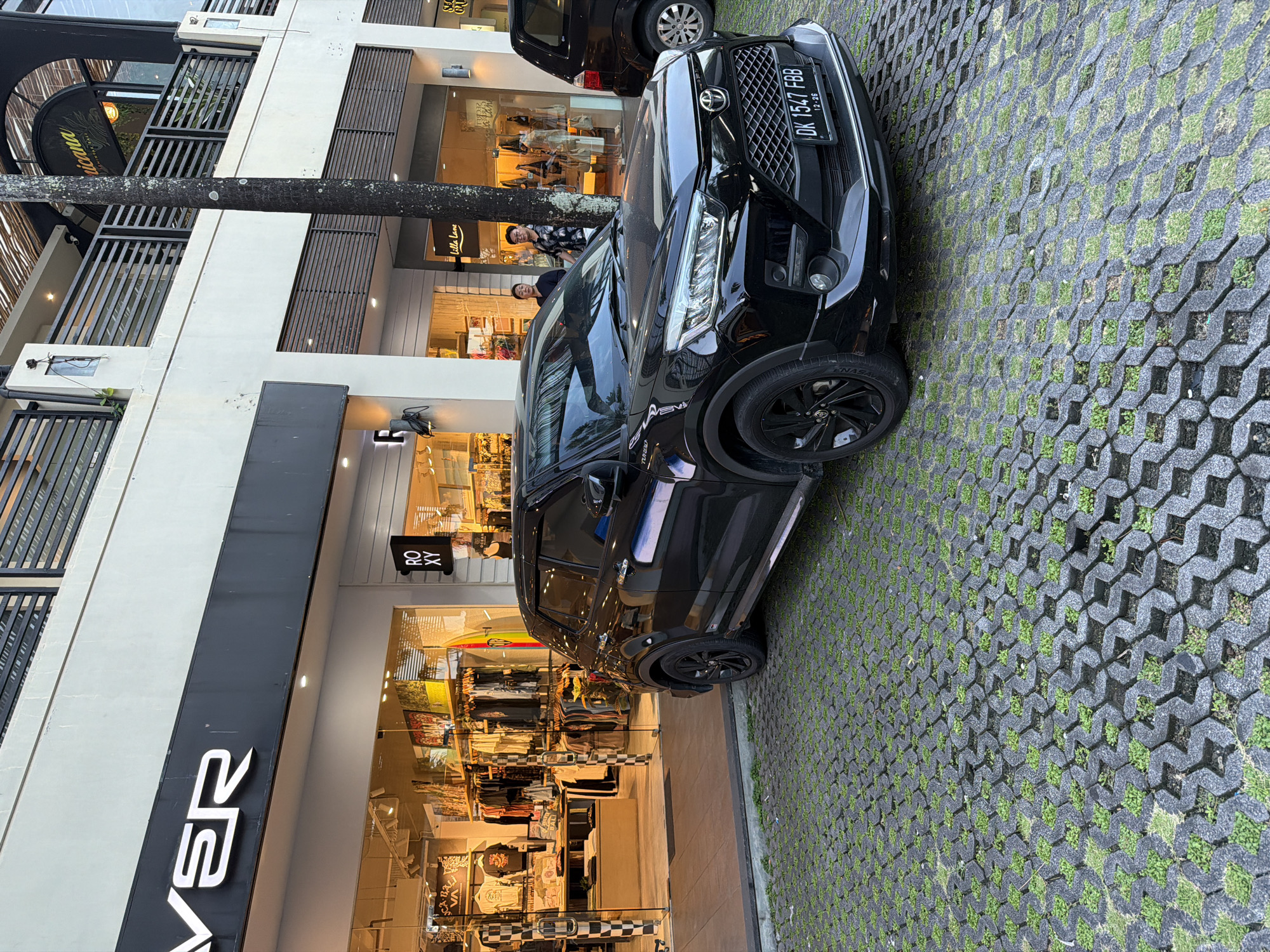 The image shows a black car parked in front of several stores in Kuta, Indonesia. The car is a Toyota, as indicated by the logo on the front grill, and its license plate reads "DK 1547 FBB". Behind the car, two people are visible in front of a store called "Villa Lena". The stores in the background are brightly lit, showcasing clothing and other items inside. The sidewalk is made of concrete blocks with grass growing in between. There's a tree trunk obstructing the front of the stores. The name of the store on the left is "VERS". A smaller sign with "ROXY" is visible near the center. Another black car is slightly visible behind the Toyota.