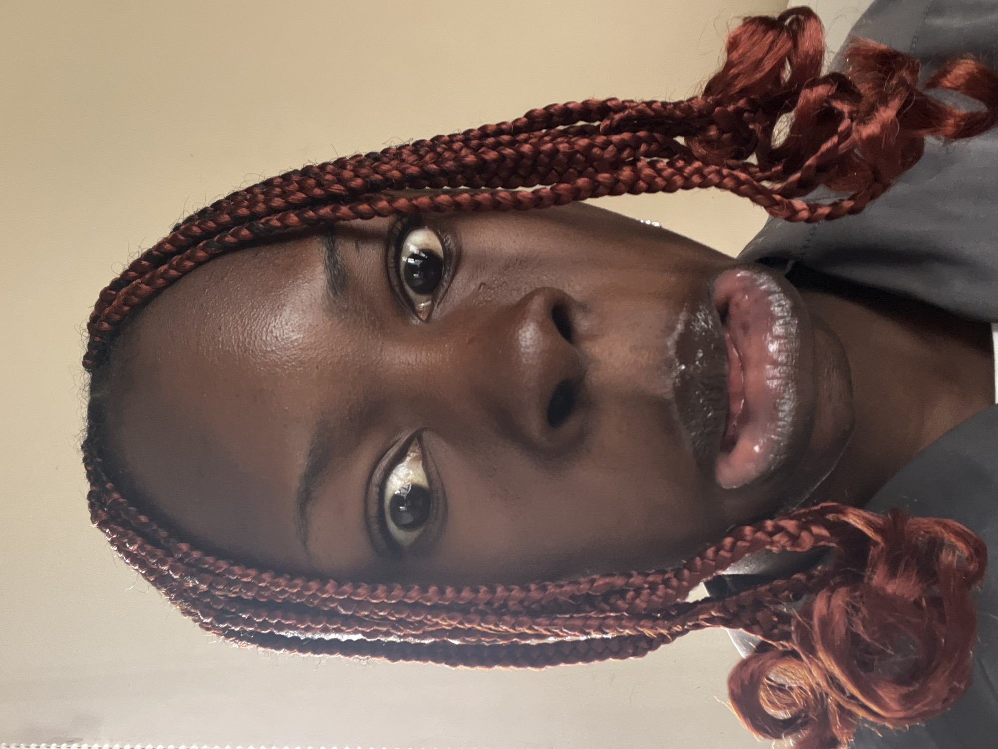 A close-up, head-on, waist-up shot shows a young Black woman with reddish-brown braided hair. Her eyes are wide open, looking slightly off to the left. Her mouth is open, with her tongue visible and her lips appearing moist and possibly glossy. She is wearing a dark gray top. The background is a plain, light beige wall. The lighting is bright and direct, casting some reflections on her face. The image is sharp and detailed, suggesting it was taken with a modern smartphone camera. There are no visible indicators of the specific location within Jos, Nigeria; however, the indoor setting with a plain wall is common in many urban environments.