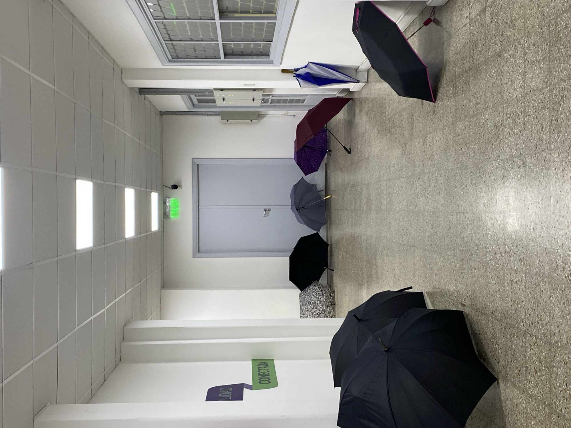 The photograph depicts a hallway in an indoor space, likely a building in Buenos Aires, Argentina. The space is characterized by white walls, a tiled floor, and a suspended ceiling with linear fluorescent lighting. A series of umbrellas are arranged along the hallway floor, varying in color and design, including black, purple, and blue umbrellas. A sign on the wall reads "CONECTADA". A double door is visible in the background. No people are present.