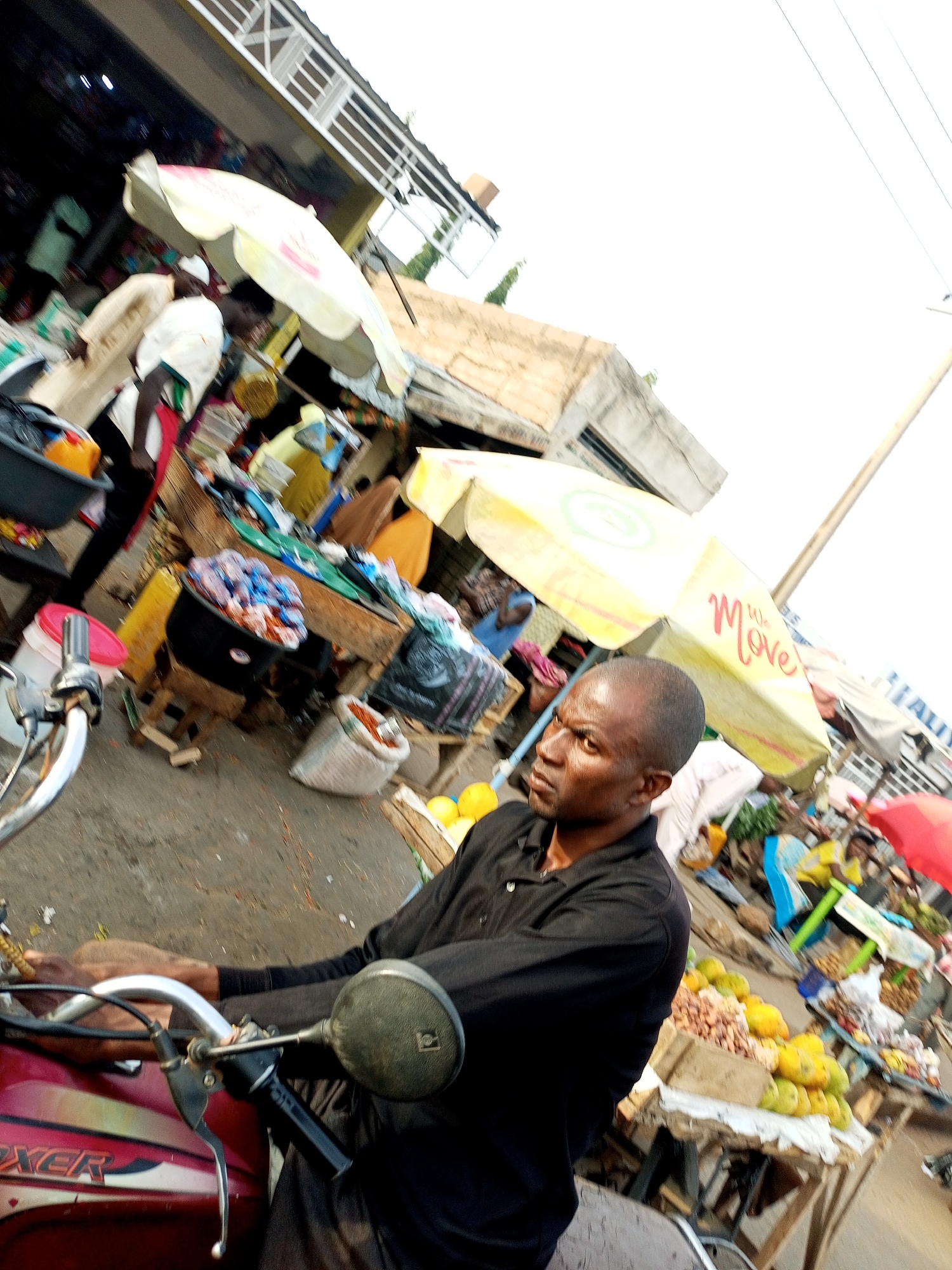 A man in a black shirt is sitting on a motorcycle, looking to his right. He is in a marketplace with several stalls and vendors. The man's face is visible, and he appears to be looking at something off-camera. Behind him, there are market stalls filled with various goods, including food items like fruits and nuts, as well as other products. There are several people in the background, some appearing to be vendors or shoppers. Several umbrellas are set up to provide shade. The scene appears to be outdoors, likely during the day, given the brightness of the sky. The overall atmosphere suggests a busy market environment. The motorcycle has the word "BOXER" visible on its side.