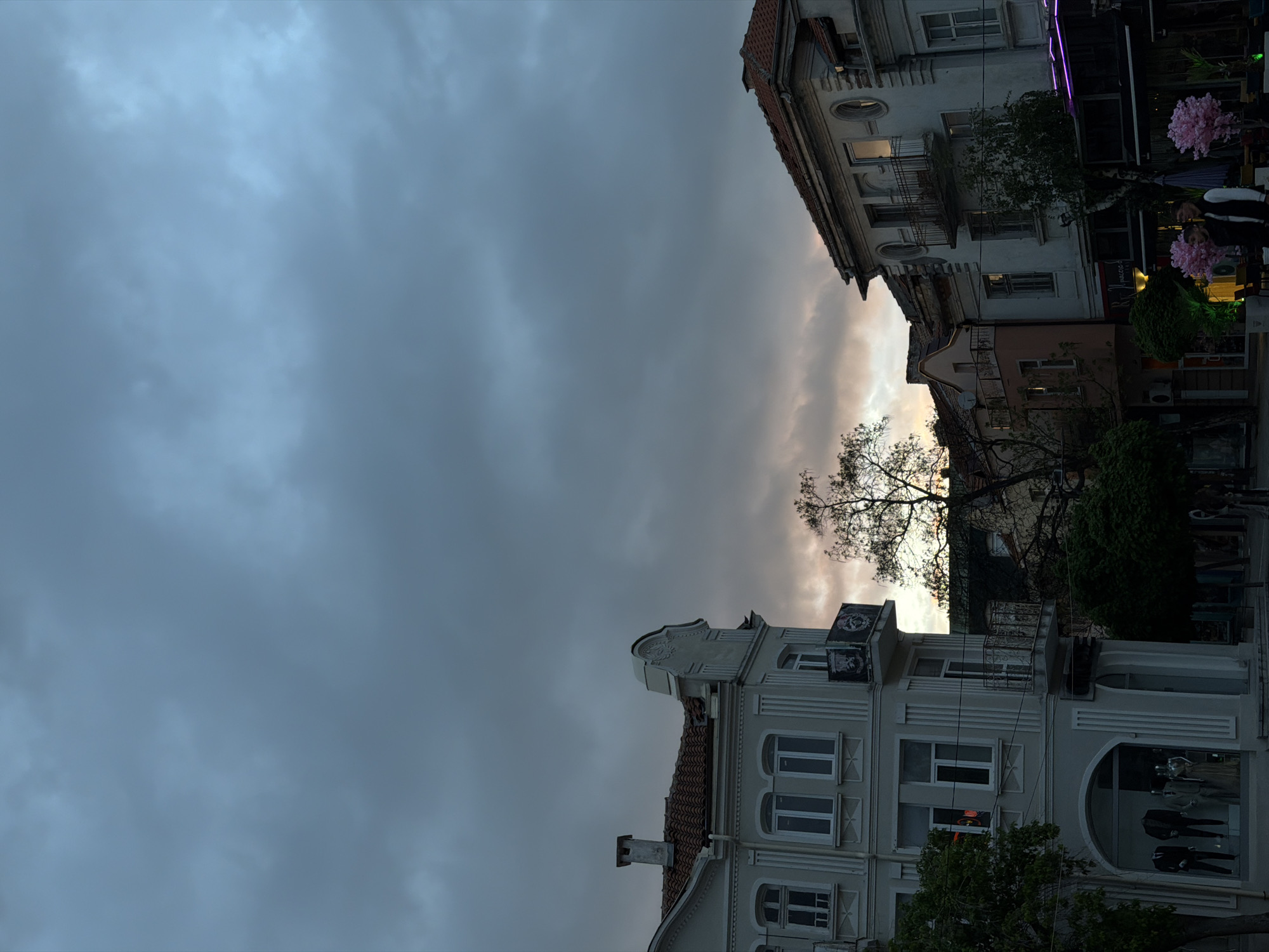 The image is of buildings in the city of Blagoevgrad, Bulgaria. The buildings are light-colored and have multiple stories. The sky is overcast with a hint of light in between the buildings. There is a tree in the center of the frame. The buildings appear to be located in an urban area, possibly a commercial district. There are mannequins visible in a shop window. People are visible outside of a building. The image is oriented vertically and appears to be taken from a low angle.