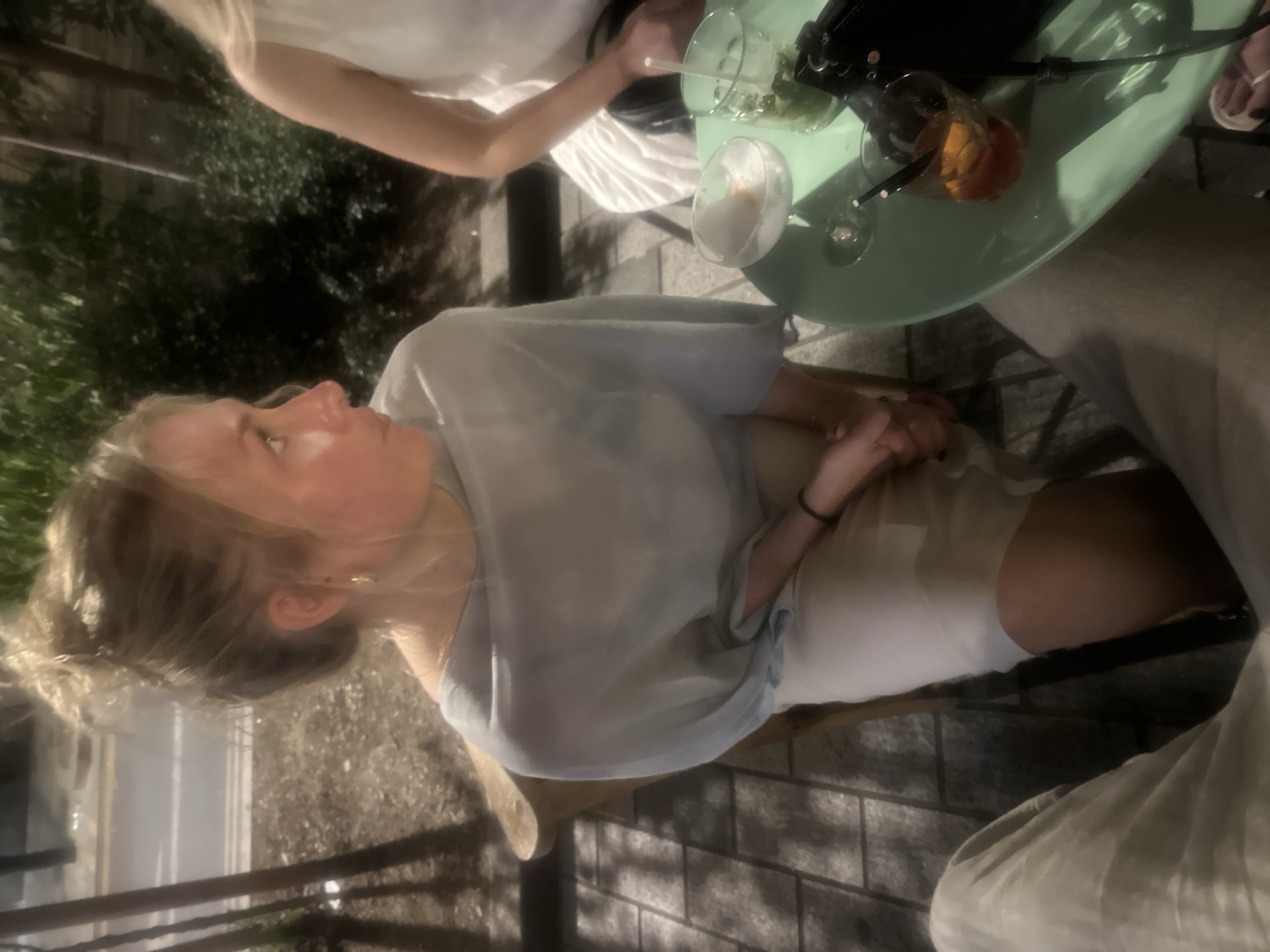 The image depicts an outdoor scene, seemingly captured in daylight. A young woman with fair skin and blonde hair pulled back in a messy bun is seated. She wears a light blue blouse over what appears to be a white dress or skirt. Her hands are clasped together in her lap.
Beside her is a circular, light green table on which several beverages sit: two clear glasses containing liquid and what appears to be ice and herbs, and a glass containing an orange-colored liquid with a garnish. Part of another person wearing a white top is visible on the left side of the table.
The setting includes an outdoor patio or sidewalk with brick or stone paving. The location is possibly in London, United Kingdom. The presence of the drinks and the casual attire suggest a relaxed social gathering.