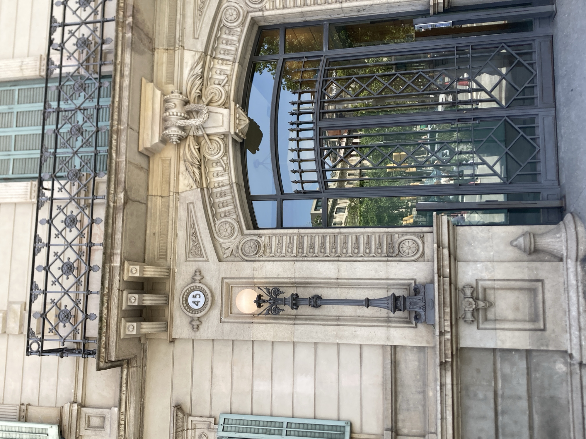 The image shows the facade of a building in Barcelona, Spain, with a prominent arched entryway. The building's architecture features stone detailing, decorative moldings, and a wrought-iron balcony above. A street lamp with an illuminated globe is mounted on the wall to the left of the entryway, near a circular plaque with the number "45". The entryway itself is a large, dark metal gate with decorative geometric patterns, set within a stone archway. The glass doors behind the gate reflect the surrounding environment, including trees, buildings, and a street with vehicles. The scene is observed from a low angle, looking upwards towards the building's entrance.