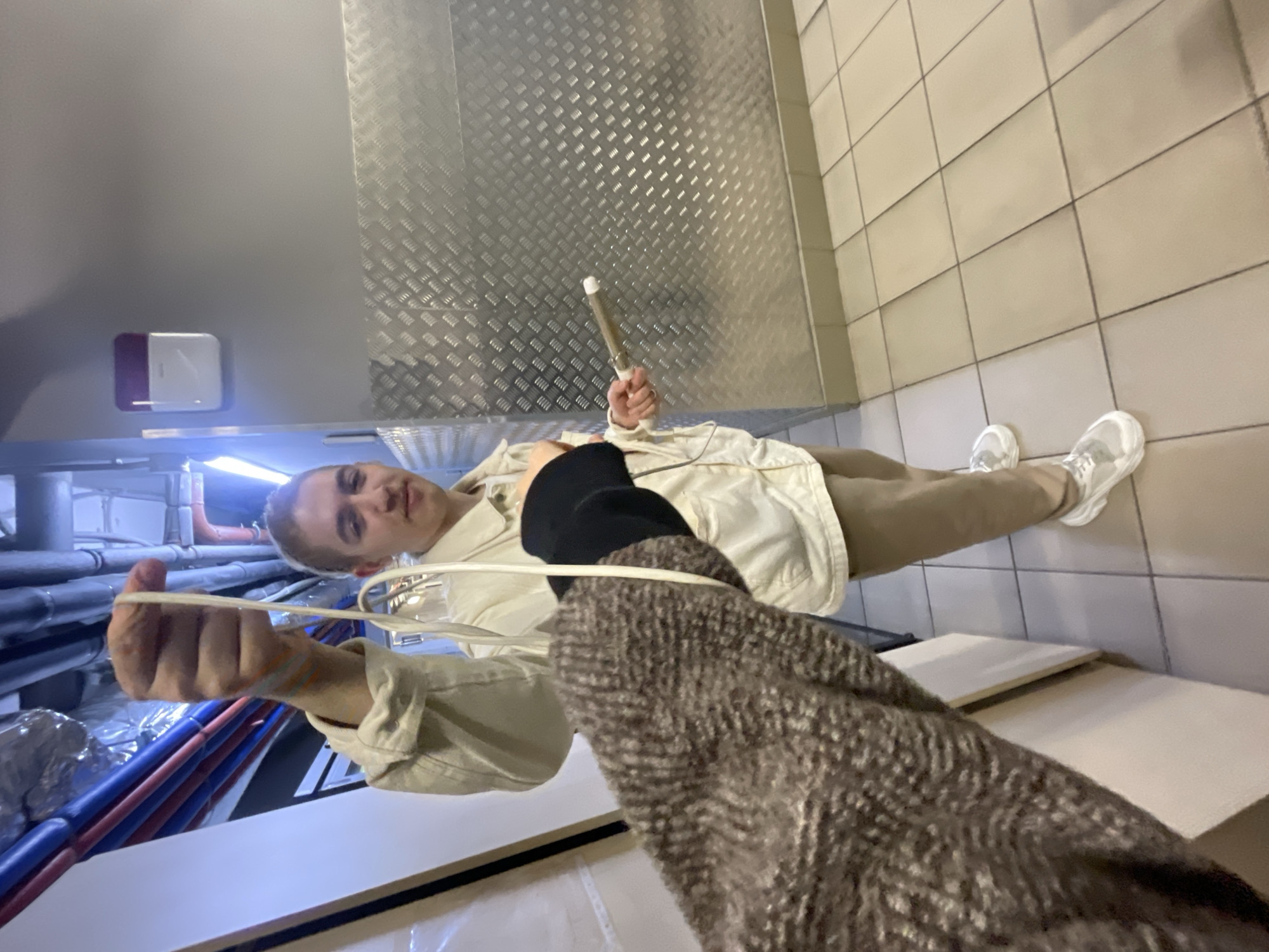 An indoor photograph, taken from a first-person perspective, captures a young person looking directly at the camera with a neutral expression. The individual, who appears to be male, has short light-colored hair and is dressed in a light-colored jacket over a black top, light pants, and white sneakers. They are holding an object resembling a hair curling wand in their left hand. Their right hand is seen stretching a light-colored elastic band or string towards the camera, interacting with the photographer whose arm, clad in dark, textured fabric, is visible in the foreground on the right.
The setting is a utilitarian or industrial interior space. The background features a reflective diamond-plate metal wall on the right and a wall with light-colored rectangular tiles on the left. The floor is also tiled. In the far left background, blue and red utility pipes and silver insulation are visible under bright artificial lighting. A white and red/purple square device, possibly a fire alarm or sensor, is mounted on a plain wall section. Light-colored shelves or surfaces are present on the right side of the image. No visible text is present.