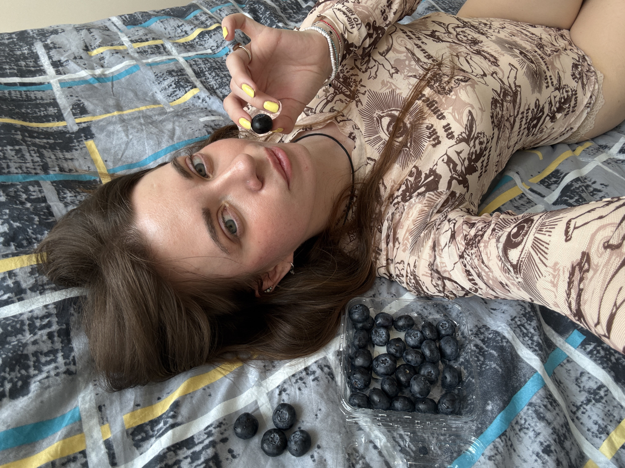 A young woman lies on a bed with a gray and blue patterned bedsheet. She is wearing a patterned tan body suit with long sleeves. In her right hand, she holds a single blueberry. A clear plastic container with additional blueberries is located near her head on the bed. The woman has brown hair, fair skin, and green eyes. Her fingernails are painted yellow. There are a few loose blueberries scattered next to the container. The location is unknown, and there is no indication of Moscow, Russia.