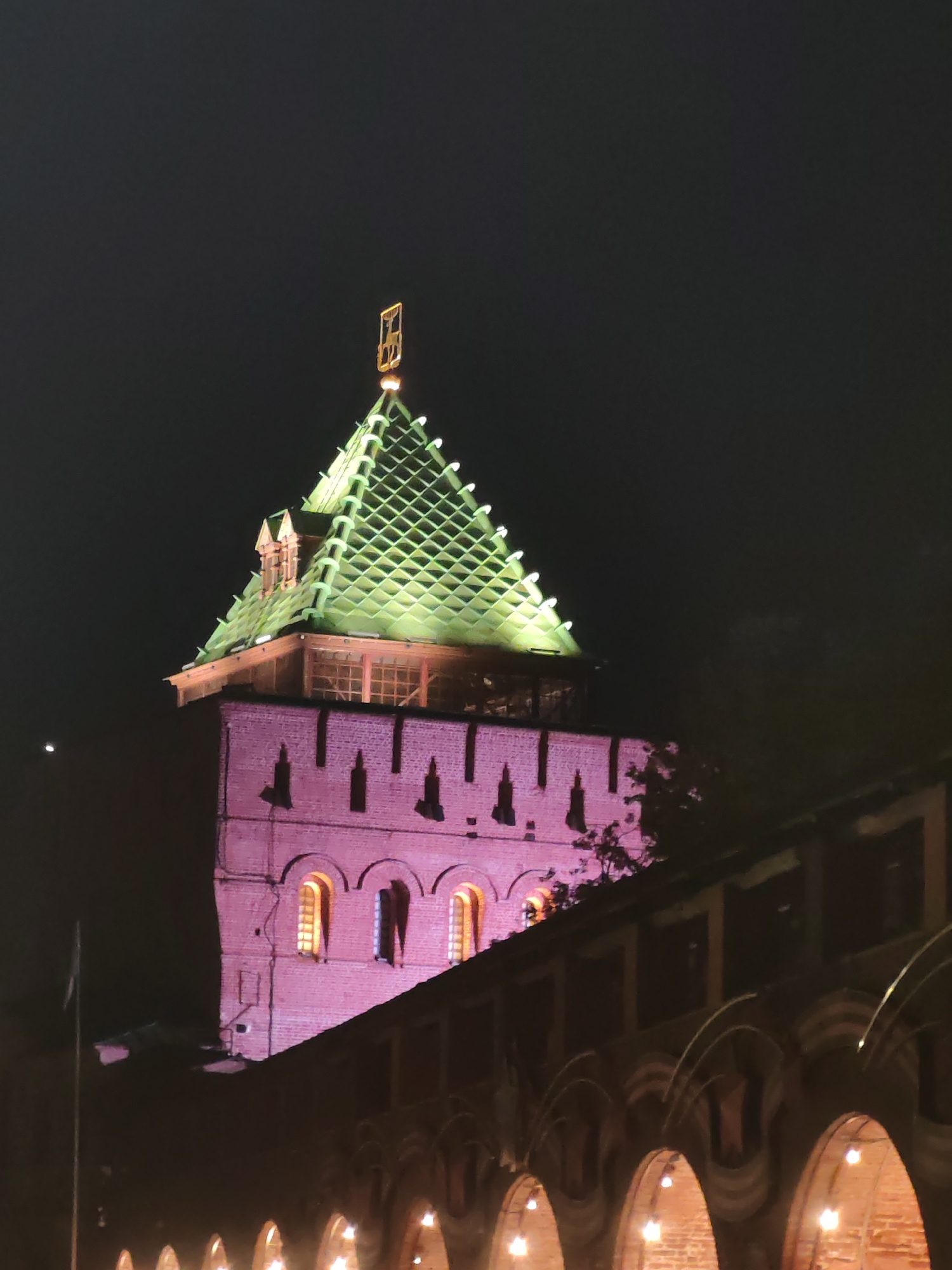 The image shows a night view of the Spasskaya Tower of the Nizhny Novgorod Kremlin. The tower is made of red brick and has a green, diamond-patterned roof topped with a golden weather vane. The tower is illuminated with pink light, and the windows are lit with yellow light. In the foreground, there is a dark, arched structure that appears to be a part of the Kremlin's fortifications, also lit with warm light. The sky is dark and clear, with a faint crescent moon visible. The overall scene depicts a historical landmark illuminated at night.