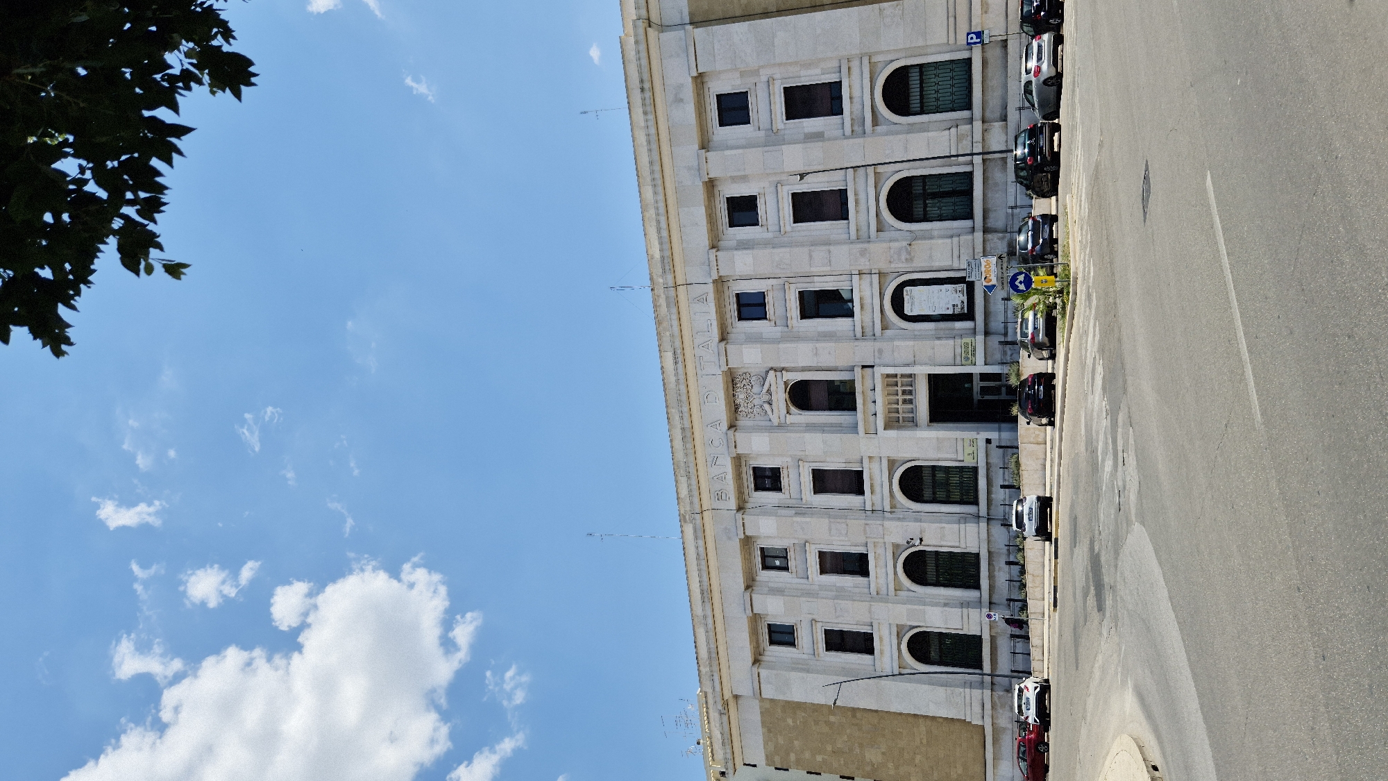 The image shows the Bank of Italy building in Taranto, Italy. It is a large, neoclassical building with a light-colored stone facade. The building is two stories tall and features a colonnade on the second floor. The words "BANCA D'ITALIA" are inscribed in large letters above the central entrance. In front of the building, there is a street with several cars parked along the sidewalk. The sky is blue with a few white clouds, and a tree branch is visible in the upper right corner of the image. The scene is bathed in sunlight, suggesting it is a clear day.