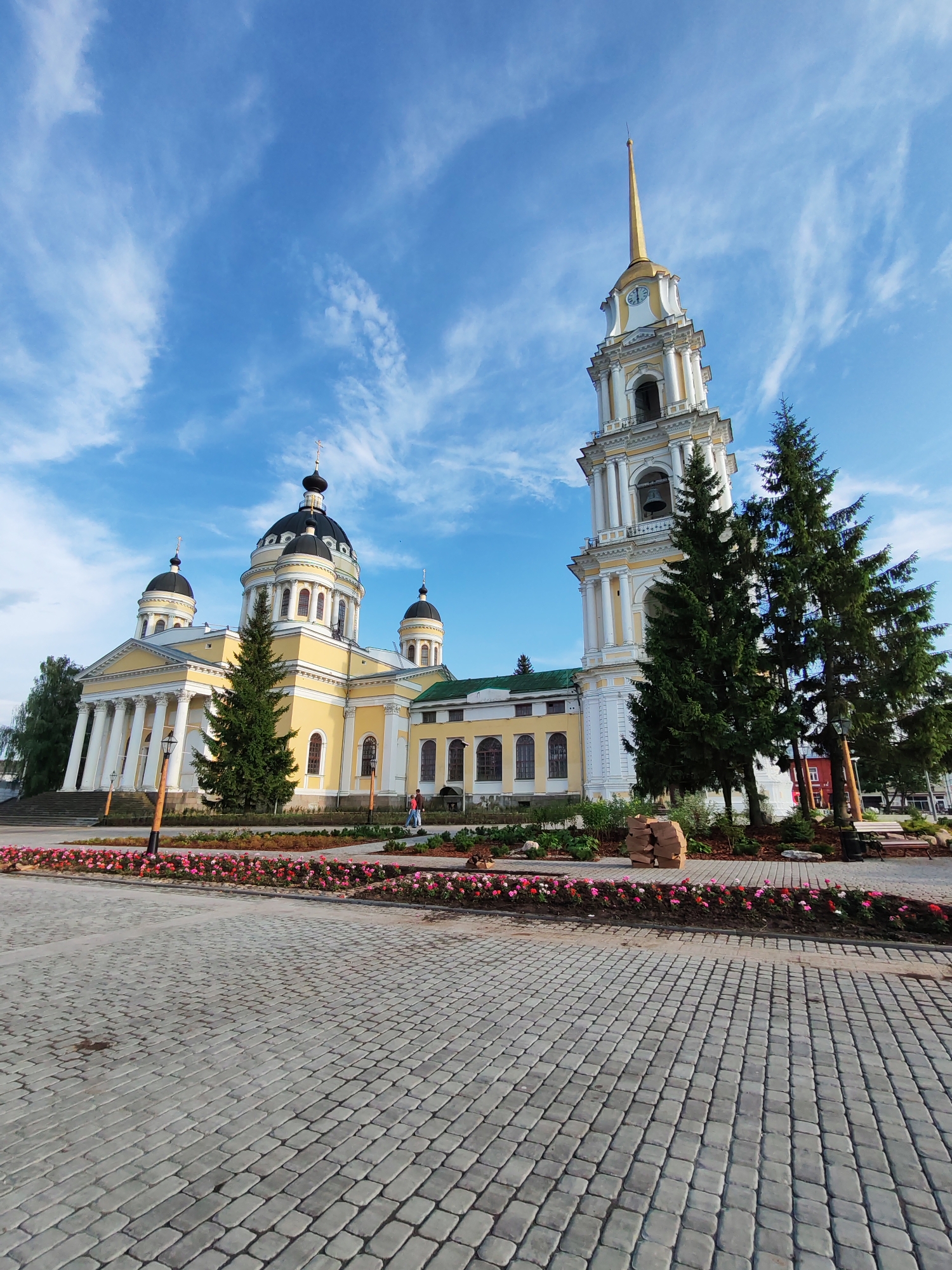 The image depicts a large, yellow Orthodox church with multiple domes, a prominent bell tower, and classical columns, situated in the city of Bol'shie Mkhi, Russia. The sky above is a vibrant blue, scattered with white, wispy clouds. In the foreground, a paved courtyard or plaza is visible, made of grey interlocking stones. A manicured flower bed with pink and red flowers lines the edge of the courtyard, separating it from a green lawn and several tall evergreen trees. Two individuals are walking in the distance on the left side of the church, near the entrance. The bell tower is tall and slender, featuring a golden spire and a clock face. The church itself is grand, with arched windows and dark domes topped with crosses. The overall scene conveys a sense of religious and civic architecture under a bright, partly cloudy sky.