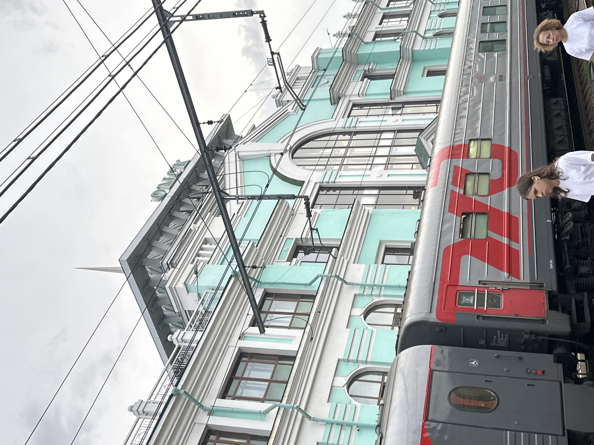 The image shows a train at the Omsk-Passazhirsky railway station in Omsk, Russia. The station building, with its distinctive turquoise and white facade and prominent "ОМСК" lettering, is visible in the background. A gray train with red accents and the Russian Railways logo is parked on the tracks. Two individuals, a young person with long hair and an older woman, stand on the platform. The sky is overcast.